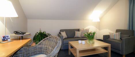 Un salon moderne avec un canapé, un fauteuil, une table basse et une chaise en osier. Il y a une lampe sur le bureau et une plante sur le sol.