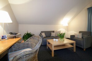 A modern living room with a sofa, armchair, coffee table, and a wicker chair. There is a lamp on the desk and a plant on the floor. - Golfhotel Fahrenbach (Trostau)