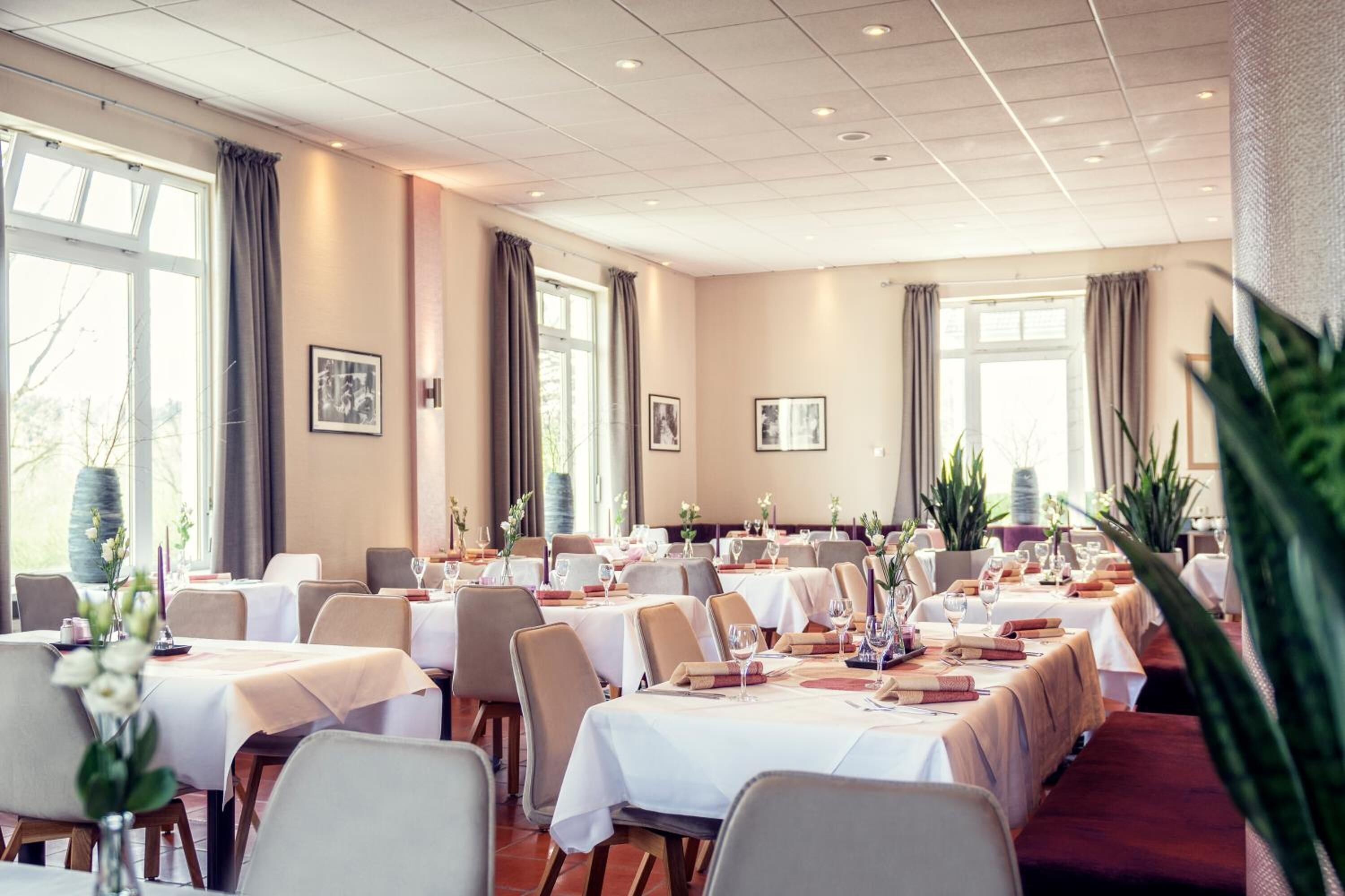 A hotel dining area with multiple tables set for dining, large windows with curtains, and indoor plants.