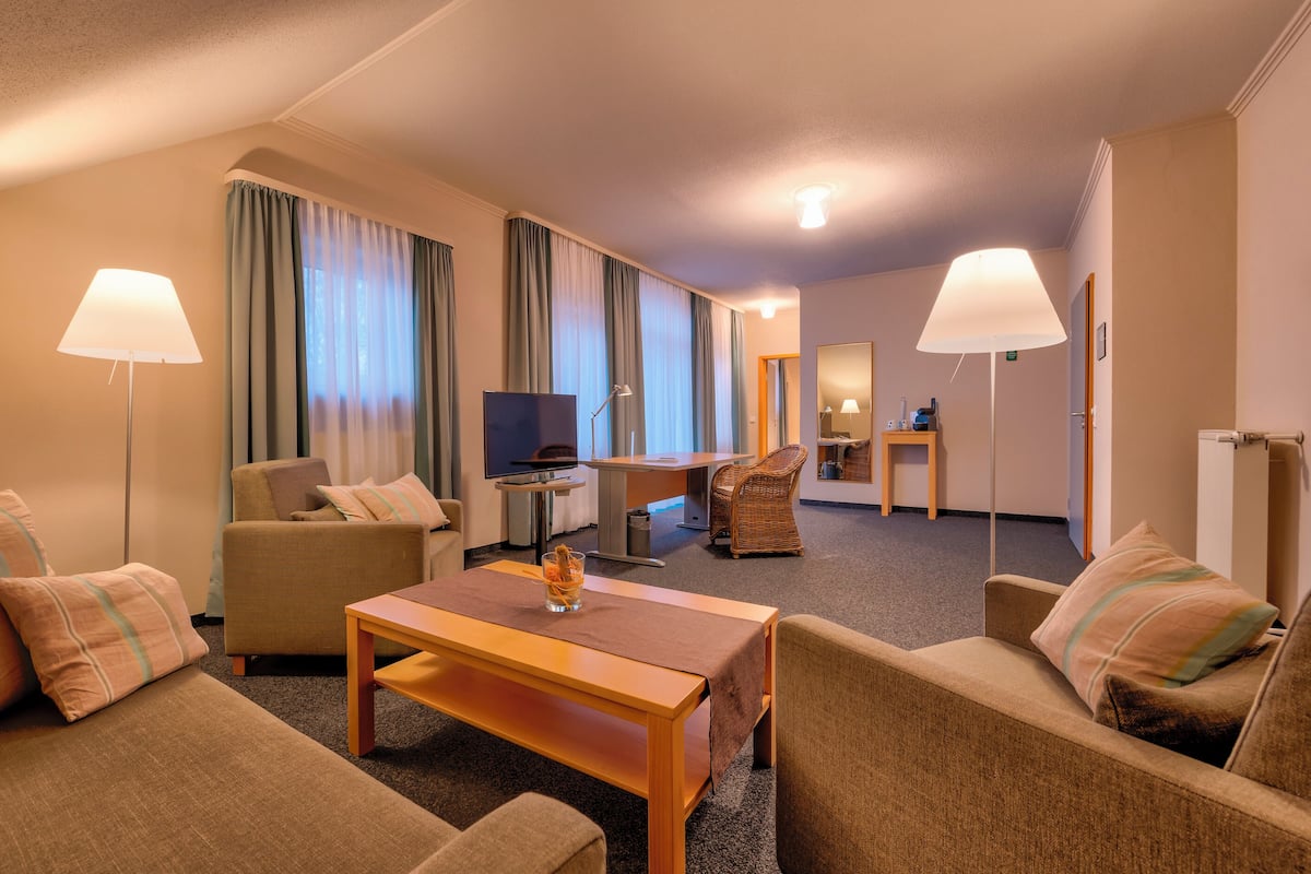 A hotel room with a sofa, armchair, and a small table. There is a TV, a lamp, and a mirror on the wall.