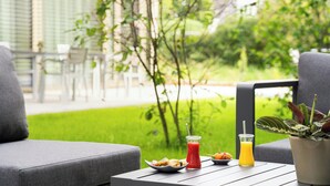 Eine Terrasse mit Sitzgelegenheiten im Freien, ein Tisch mit Speisen und Getränken und Blick auf eine grüne Rasenfläche und Bäume.
