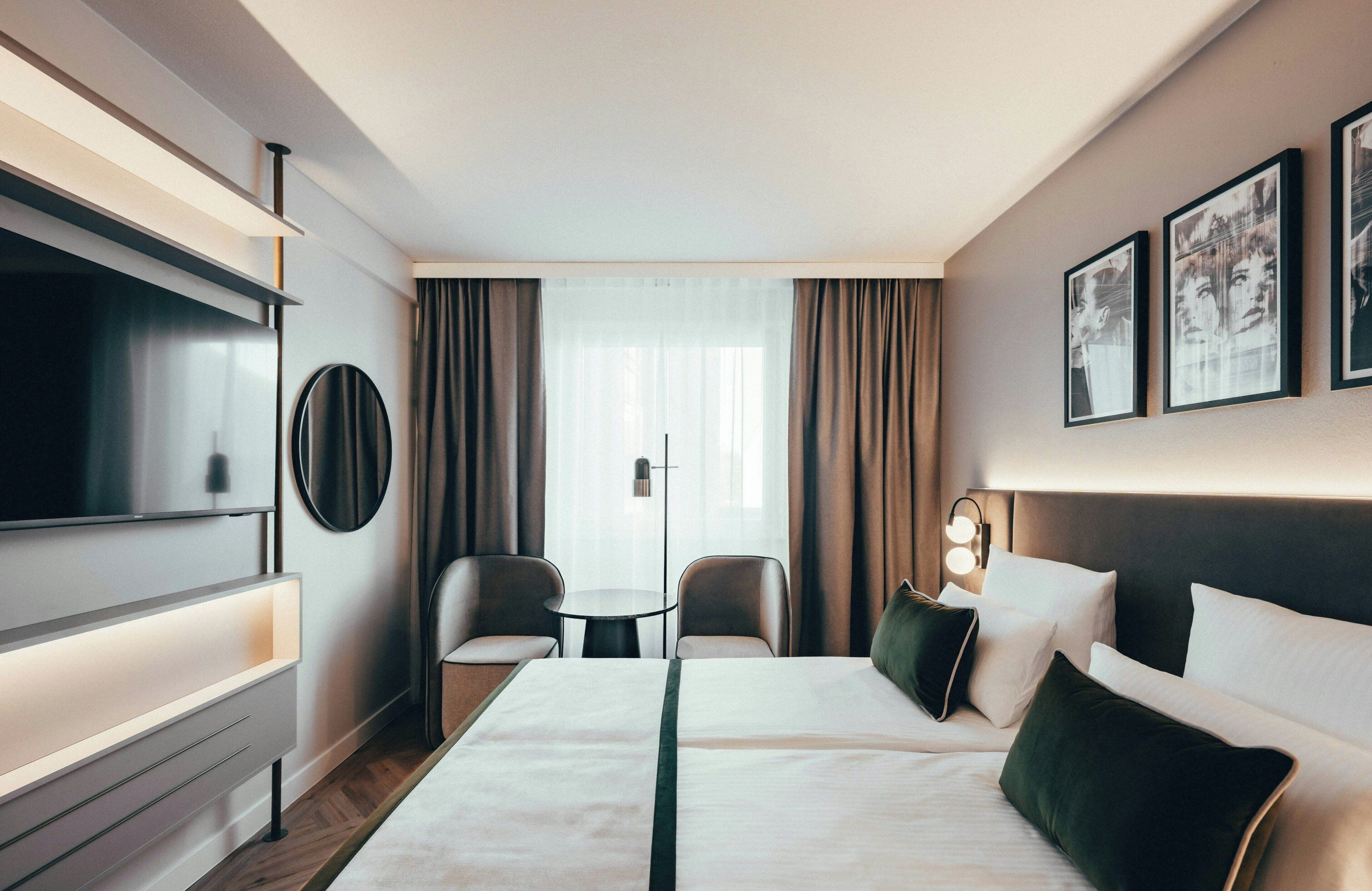 A hotel room with a bed, two chairs, a TV, a small table, and a wall-mounted light.