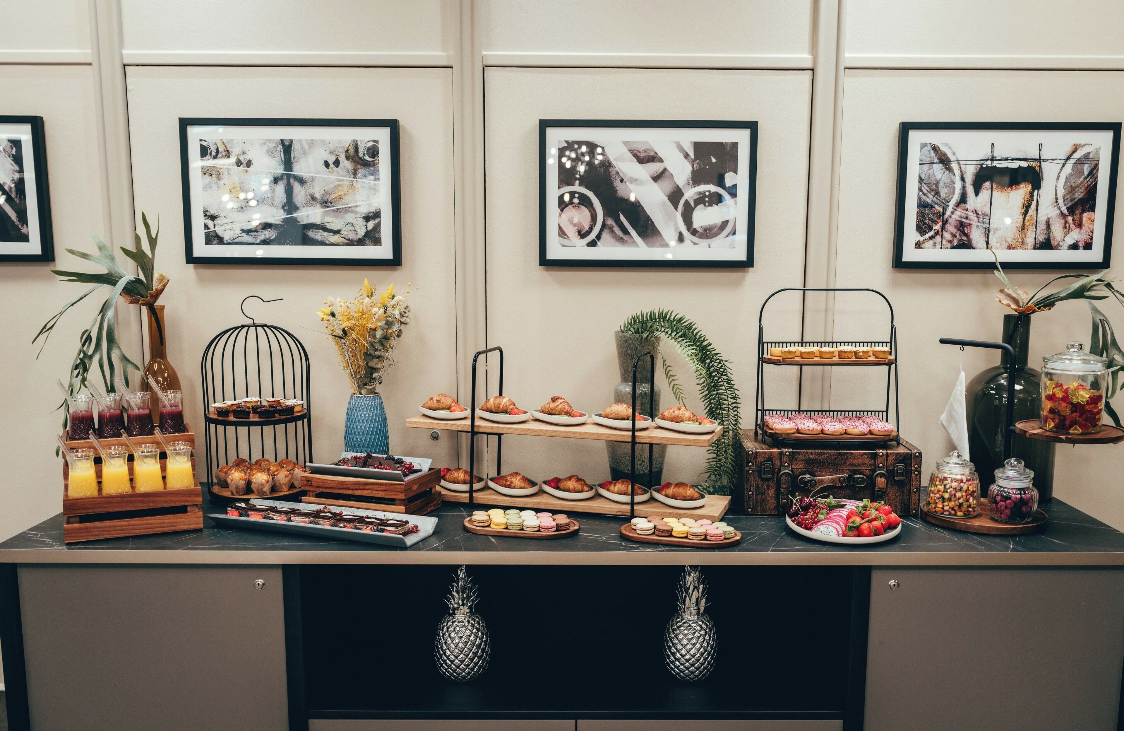 A buffet setup with various food items and drinks, framed artwork on the wall, and a decorative plant.