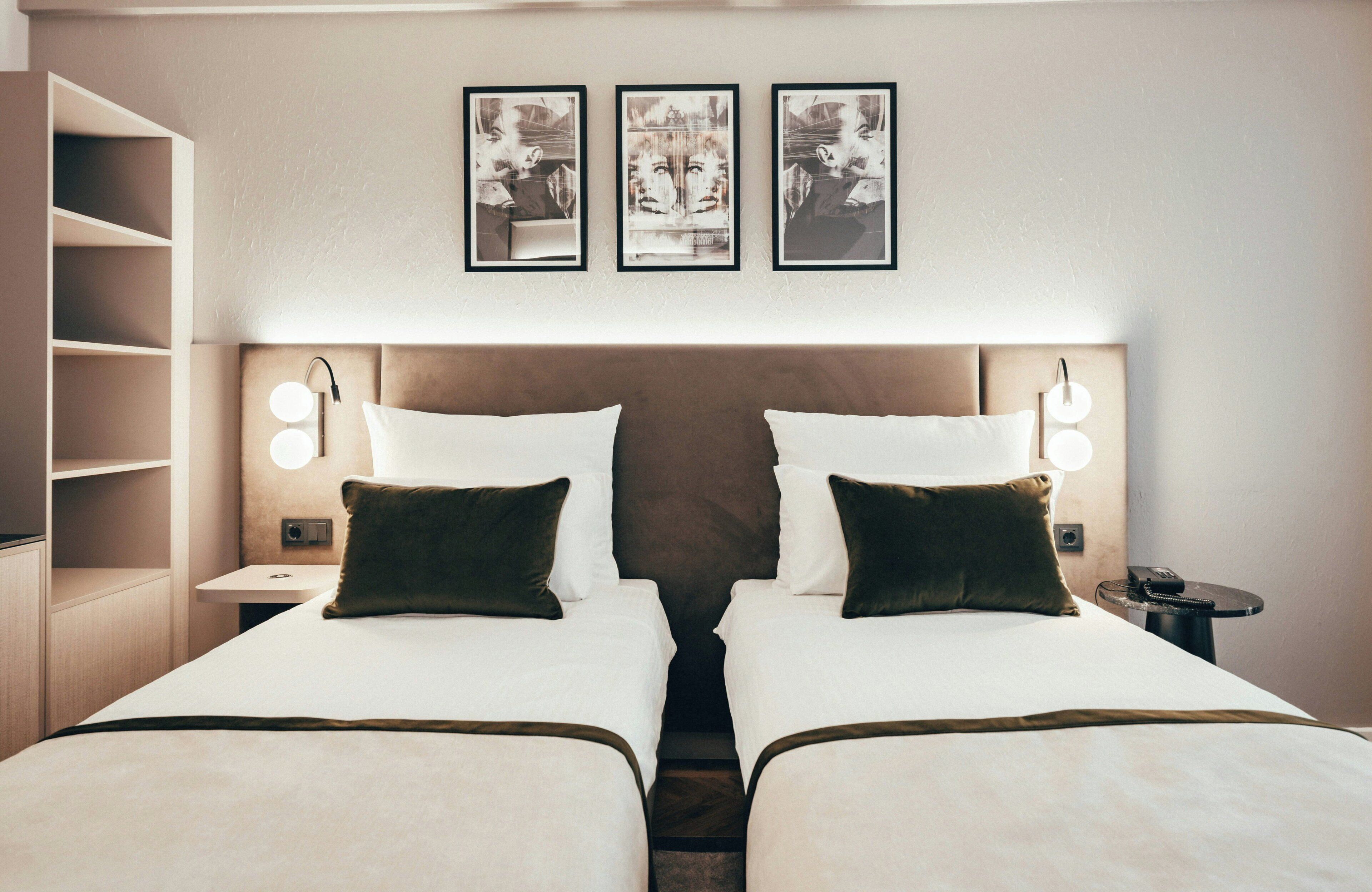 Ein Hotelzimmer mit zwei Betten, einem Nachttisch mit Lampe, einem Wandregal und drei gerahmten Bildern über den Betten.