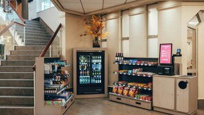 Un vestíbulo de hotel con una escalera, un refrigerador de bebidas, máquinas expendedoras de snacks y una pantalla digital.