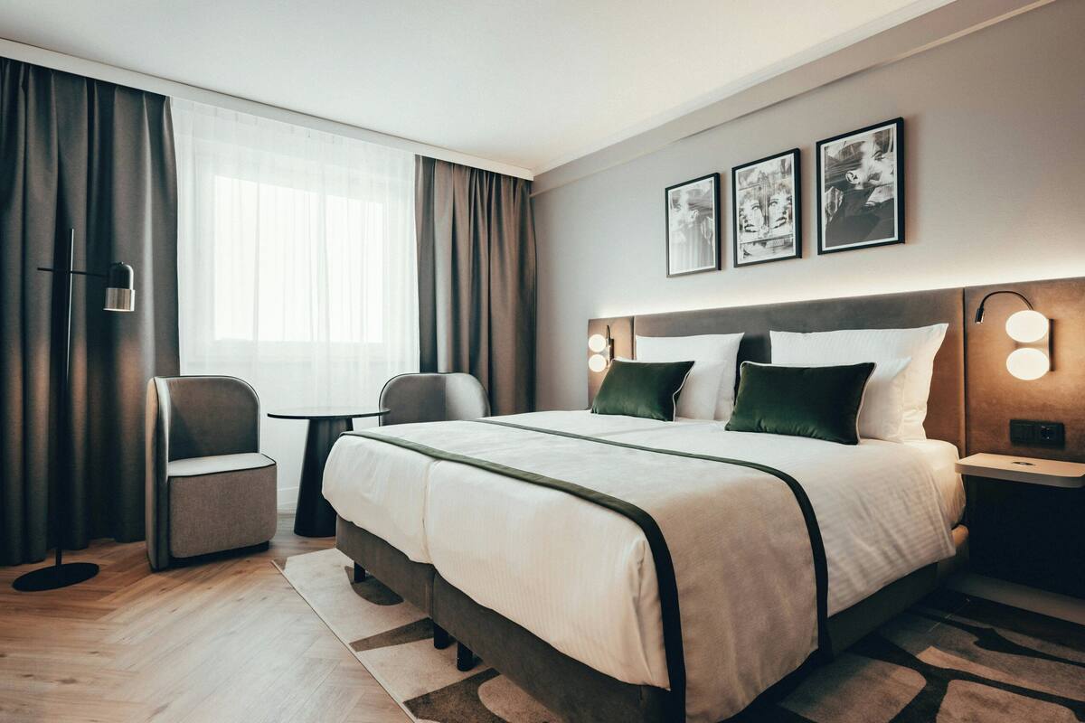 A modern hotel room with a large bed, a desk, a chair, and a window with curtains.