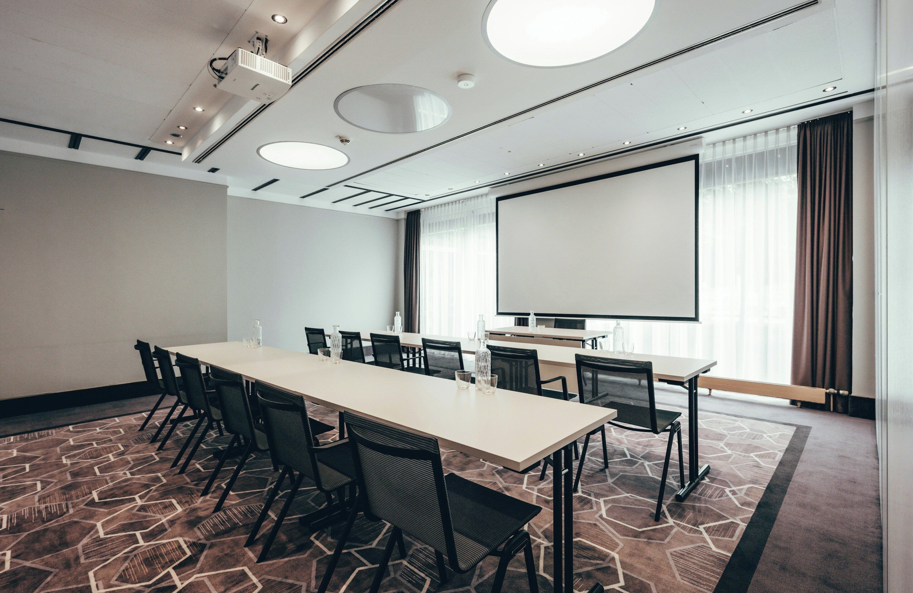 A conference room with a long table, chairs, a projector screen, and a large window with curtains.