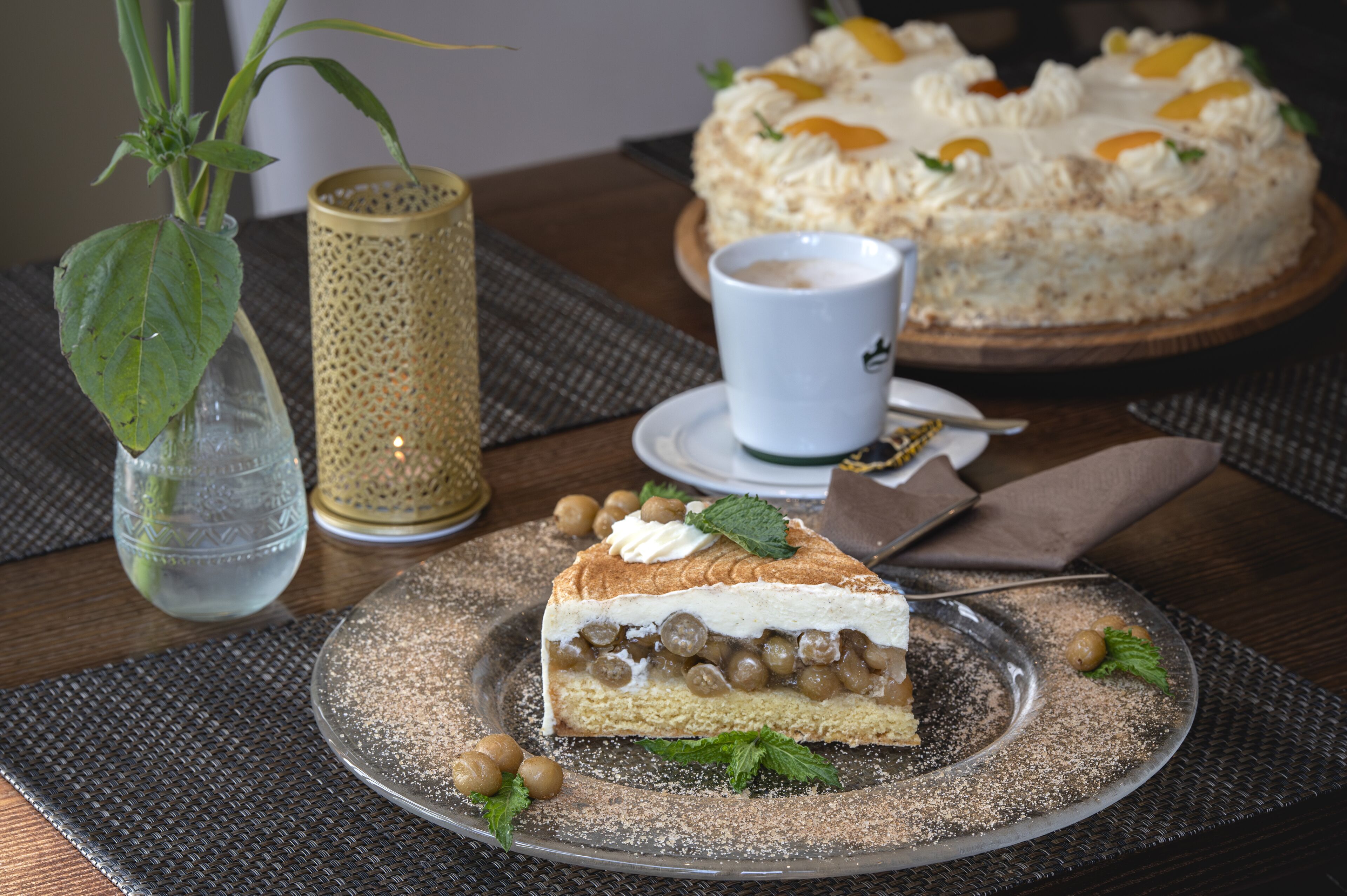 Una rebanada de pastel con una capa cremosa y cobertura de fruta, una taza de café, y un pastel entero al fondo sobre una mesa.