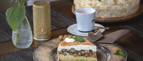 Una rebanada de pastel con una capa cremosa y cobertura de fruta, una taza de café, y un pastel entero al fondo sobre una mesa.