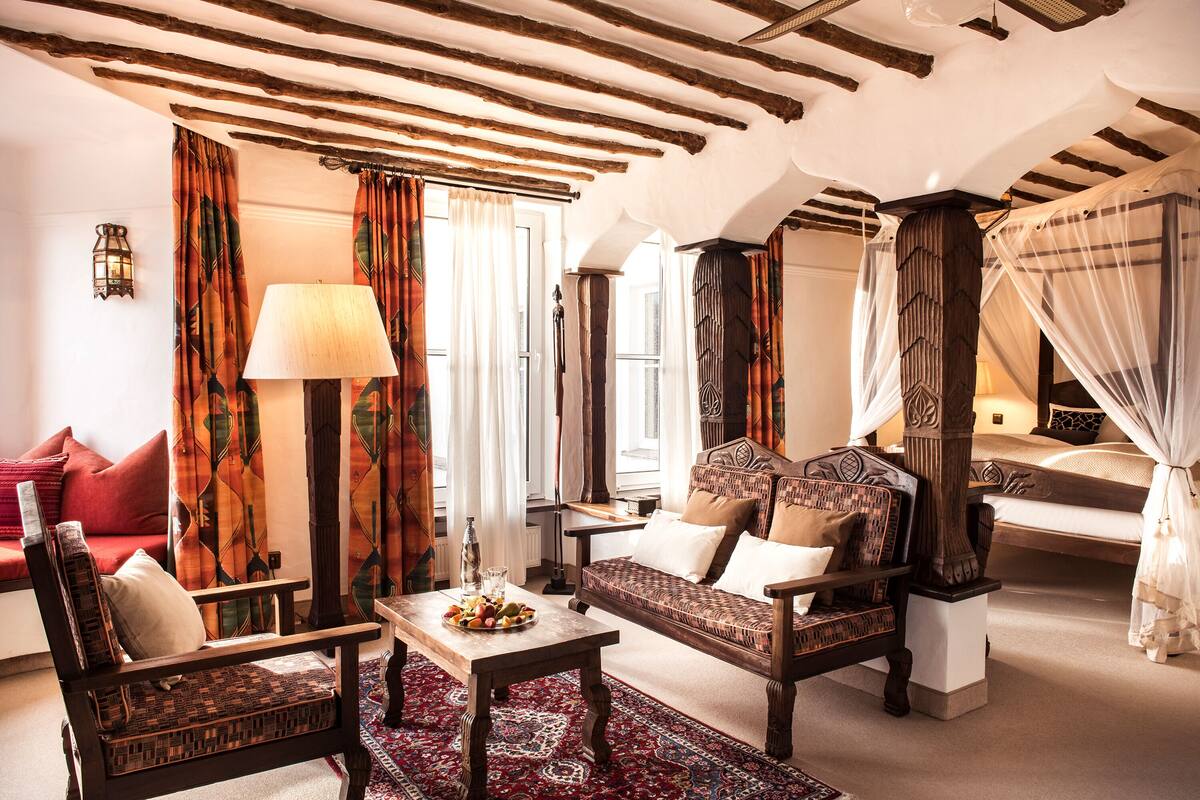 A traditional room with a sofa, armchairs, a table with fruit, and a canopy bed.