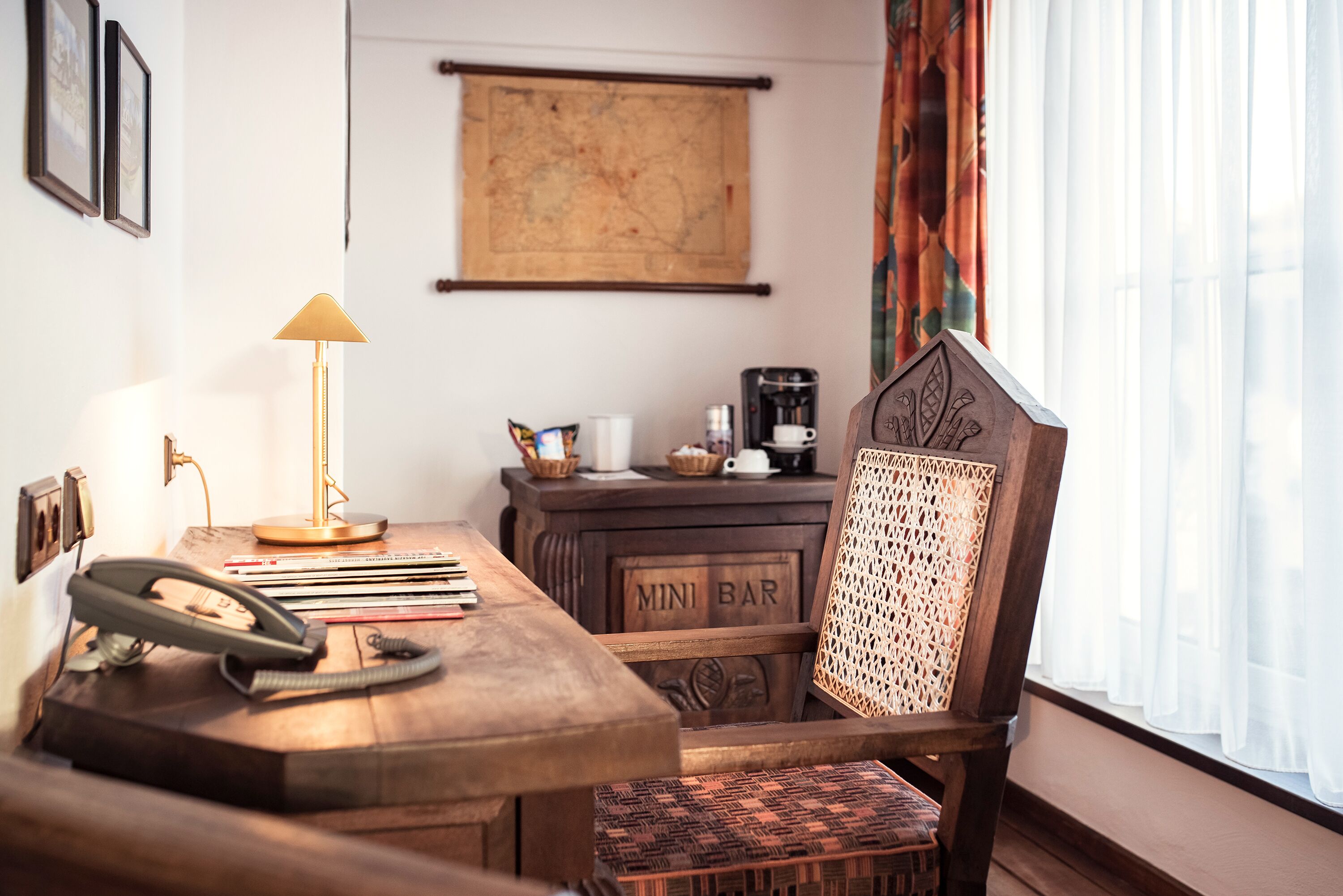 Escritorio de madera con teléfono, lámpara y cafetera. Hay una silla con respaldo tejido y un cartel que dice “MINI BAR”.