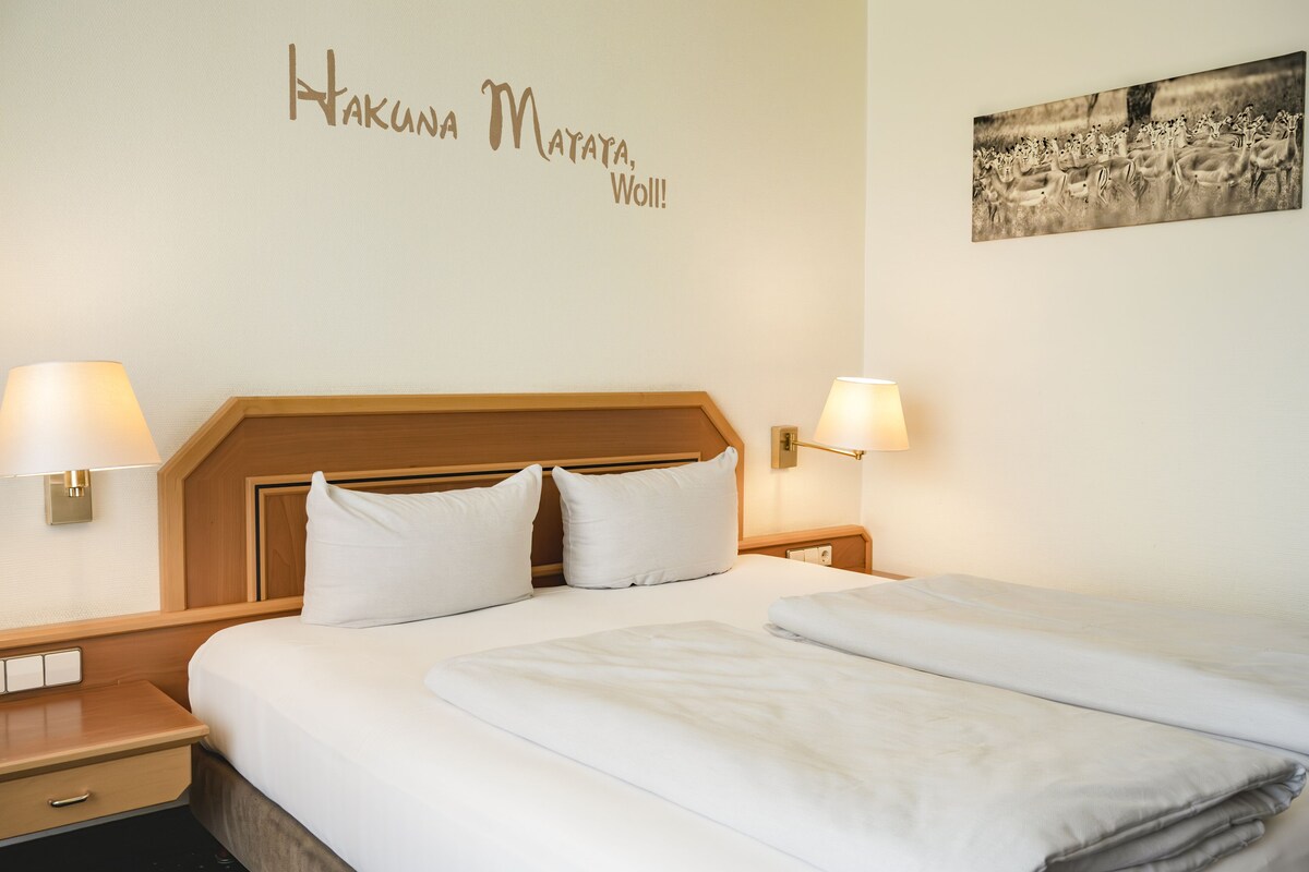 Habitación de hotel con cabecera de madera, dos lámparas de noche, cama con ropa blanca, mesita de noche con cajón y un adorno en la pared.