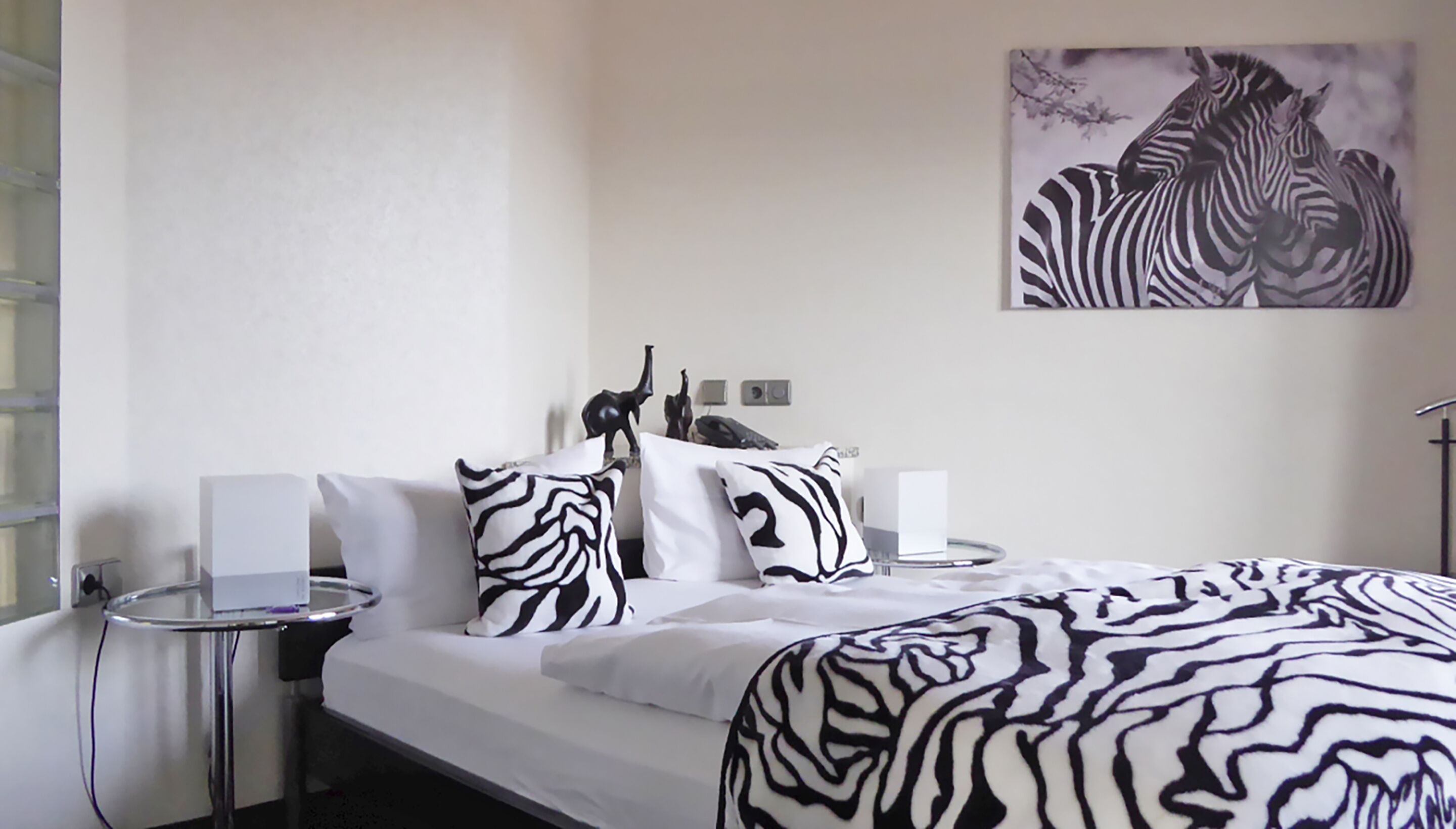 A hotel room with a bed, white bedding, zebra-patterned pillows, a zebra artwork on the wall, a small table with a lamp, and a decorative elephant statue.