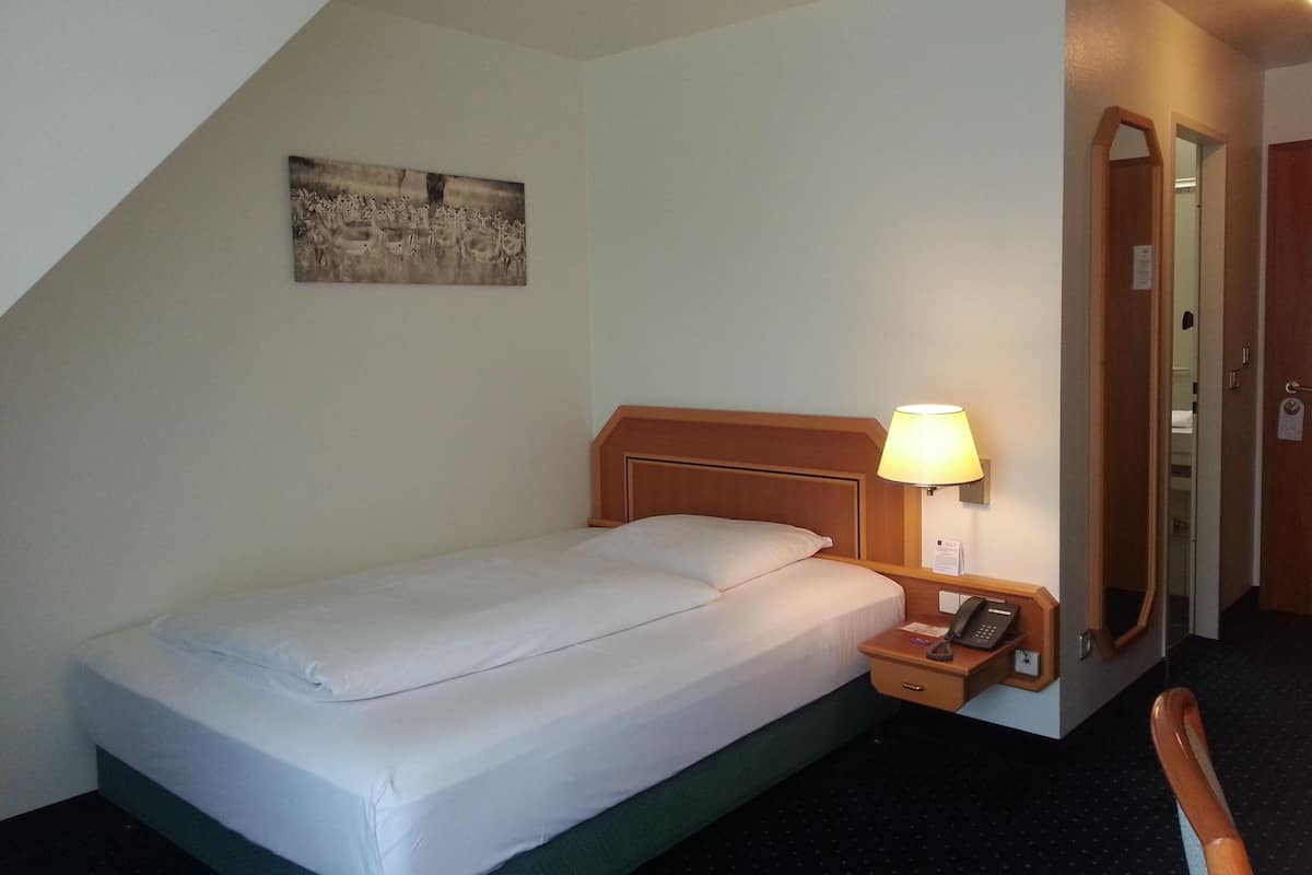 A hotel room with a single bed, a bedside table with a lamp, a telephone, a mirror, and a painting on the wall.