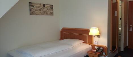A hotel room with a single bed, a bedside table with a lamp, a telephone, a mirror, and a painting on the wall.