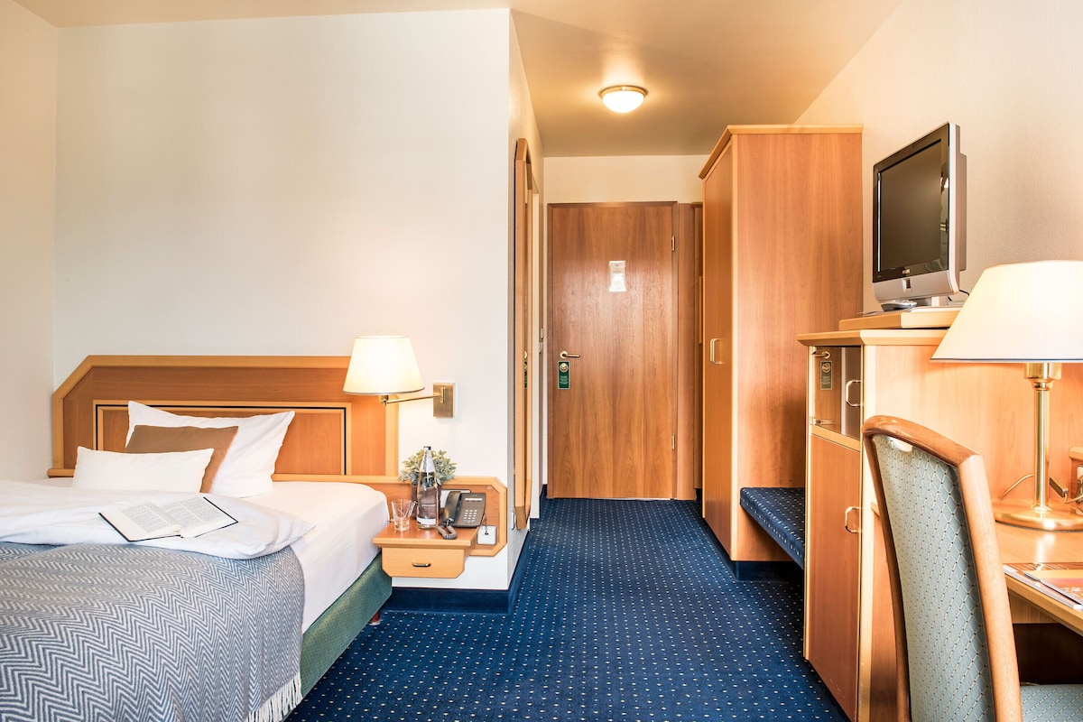 A hotel room with a bed, bedside table, lamp, TV, and a desk with a chair.