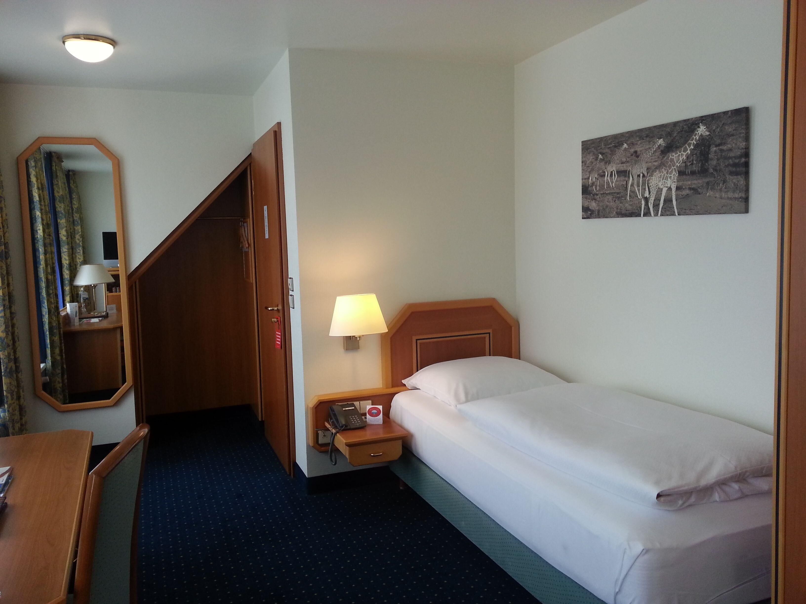 A hotel room with a bed, bedside lamp, a telephone, a mirror, a desk, and a painting on the wall.