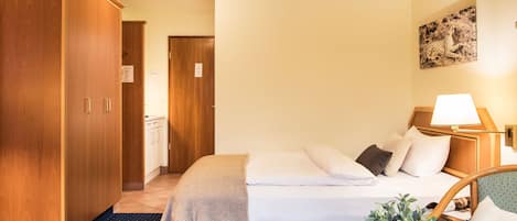 Una habitación de hotel con cama, mesita de noche con lámpara, planta en maceta y armario.