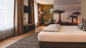Habitación de hotel con cama, escritorio, silla, lámpara y un mural de árboles.