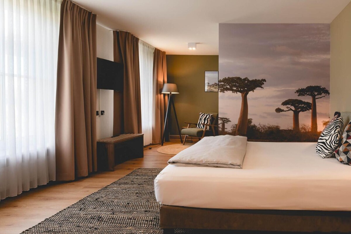 Habitación de hotel con cama, escritorio, silla, lámpara y un mural de árboles.