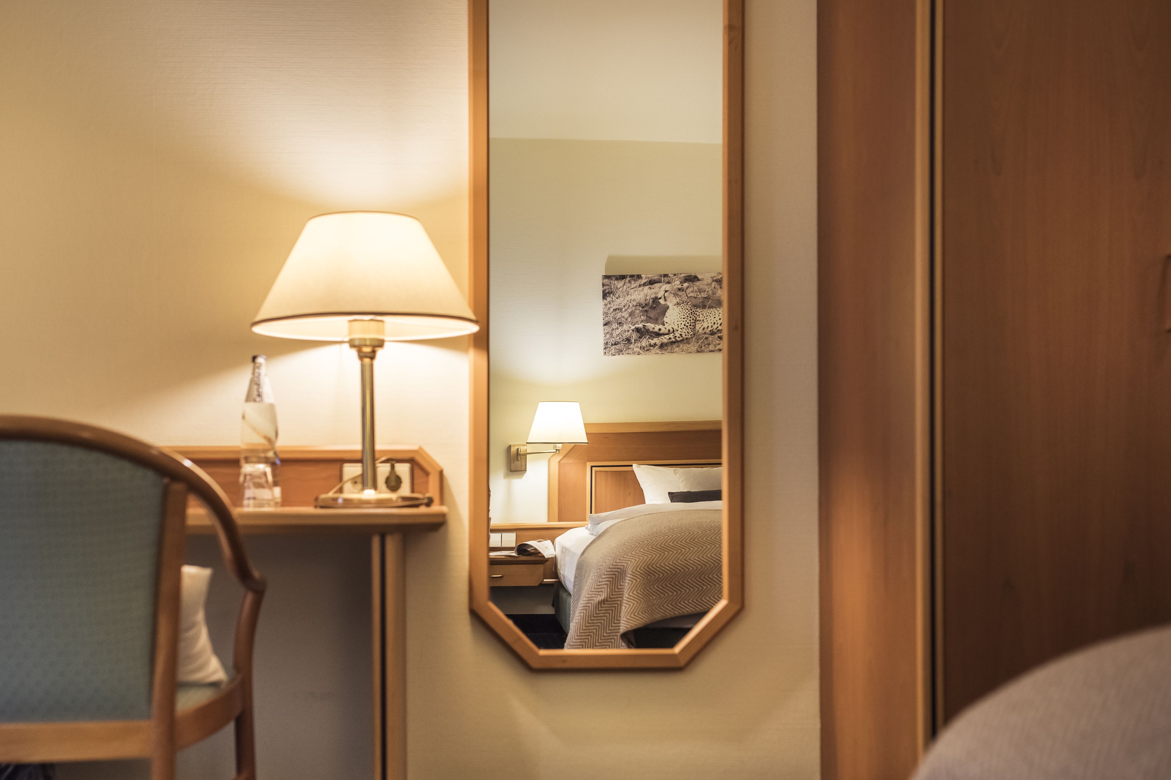 A hotel room with a bed, a nightstand, a lamp, and a mirror reflecting the room.