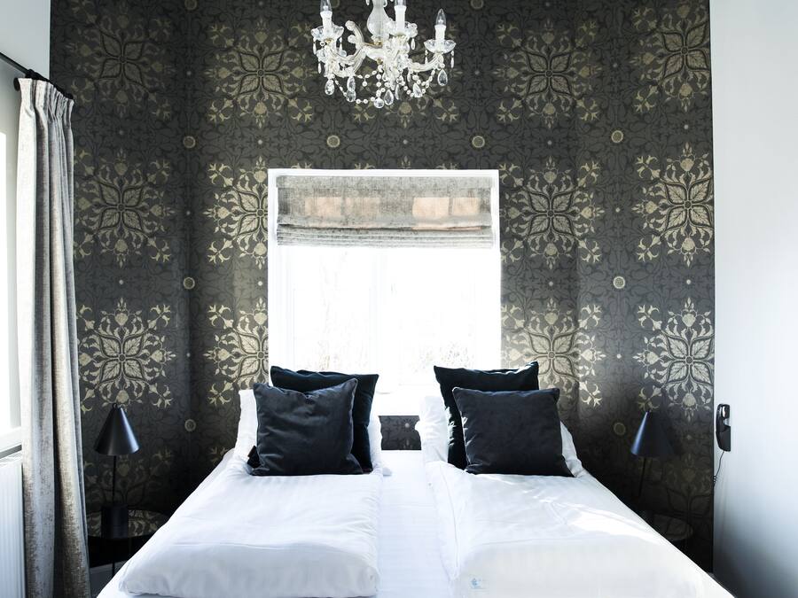 A bedroom with a large bed, dark patterned wallpaper, a chandelier, and a window.