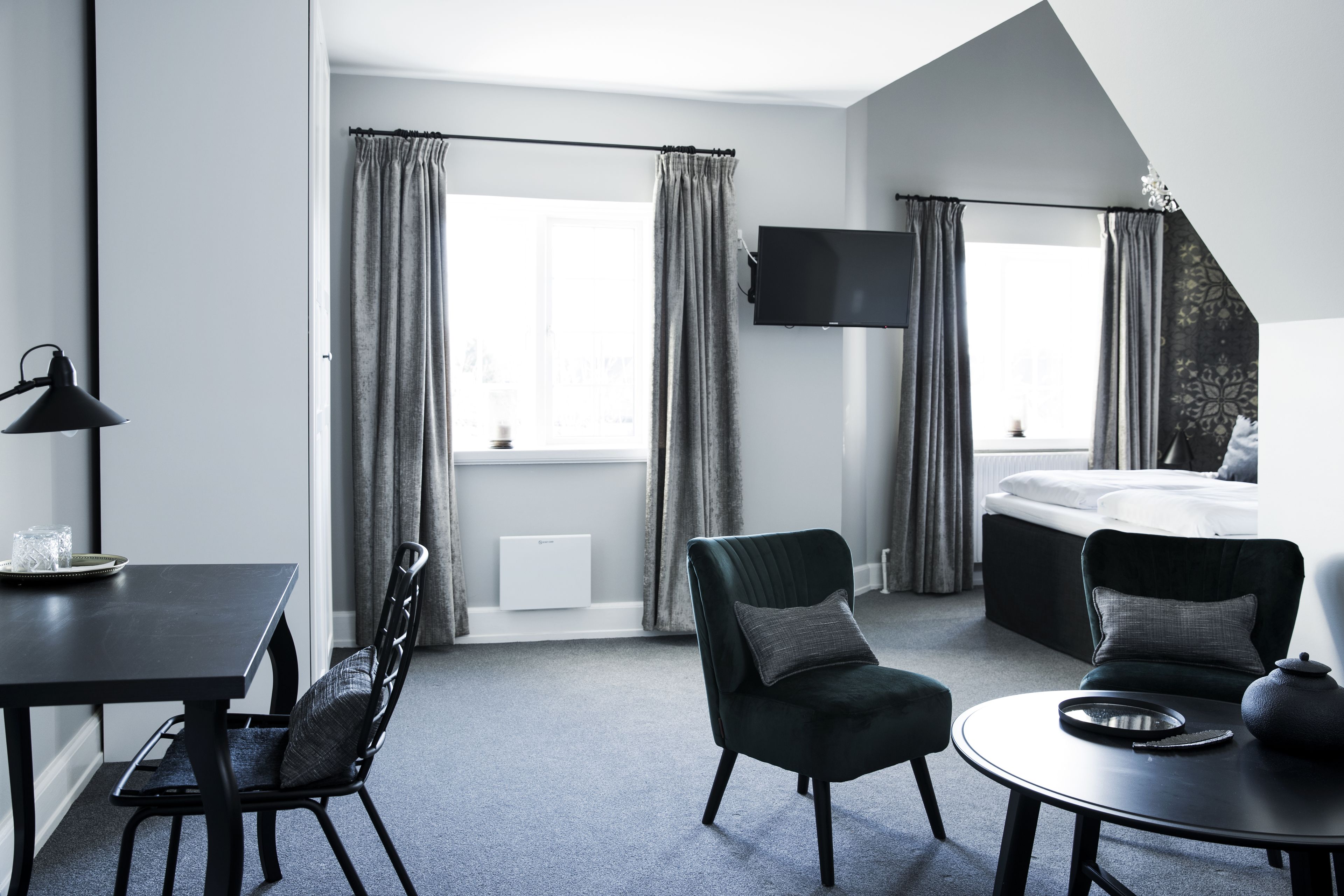 A modern hotel room with a bed, a dining table, two chairs, a TV, and large windows with curtains.
