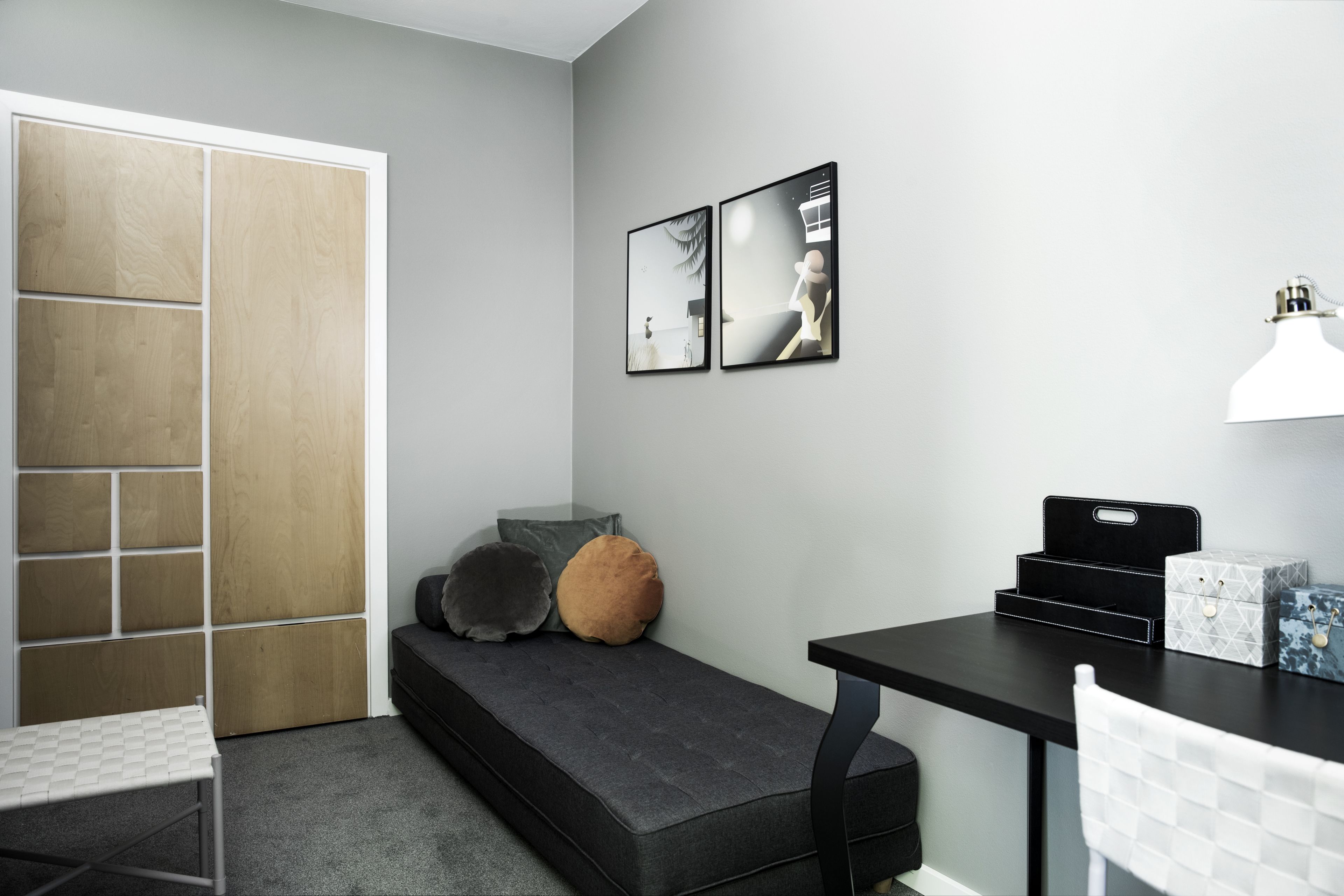 Una habitación compacta con sofá, escritorio y silla. Hay dos cuadros en la pared, una lámpara blanca en el escritorio y una mesita con cajas.
