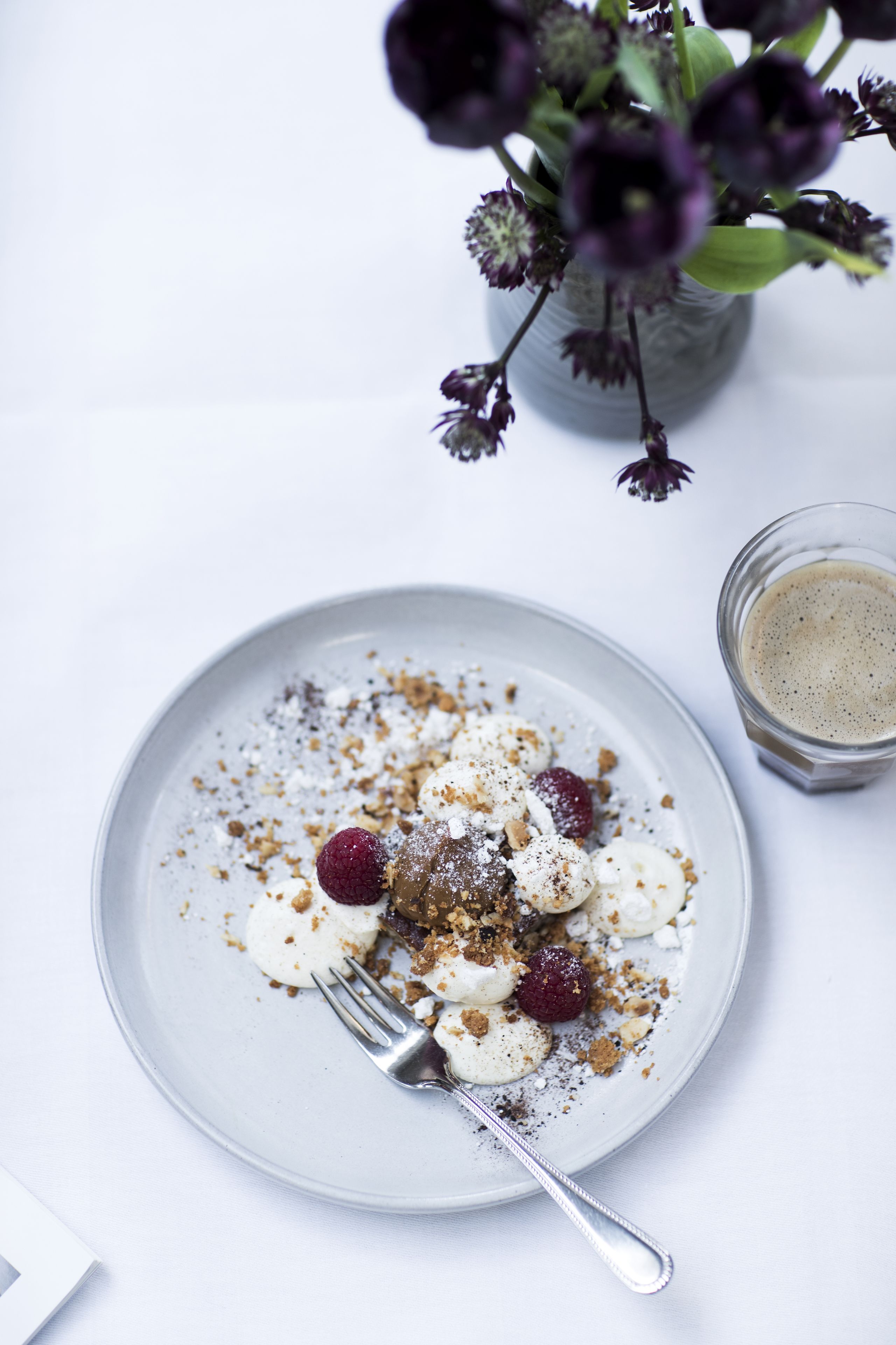 Une assiette de dessert avec des fruits rouges et une garniture émiettée, une tasse de café et un vase rempli de fleurs.