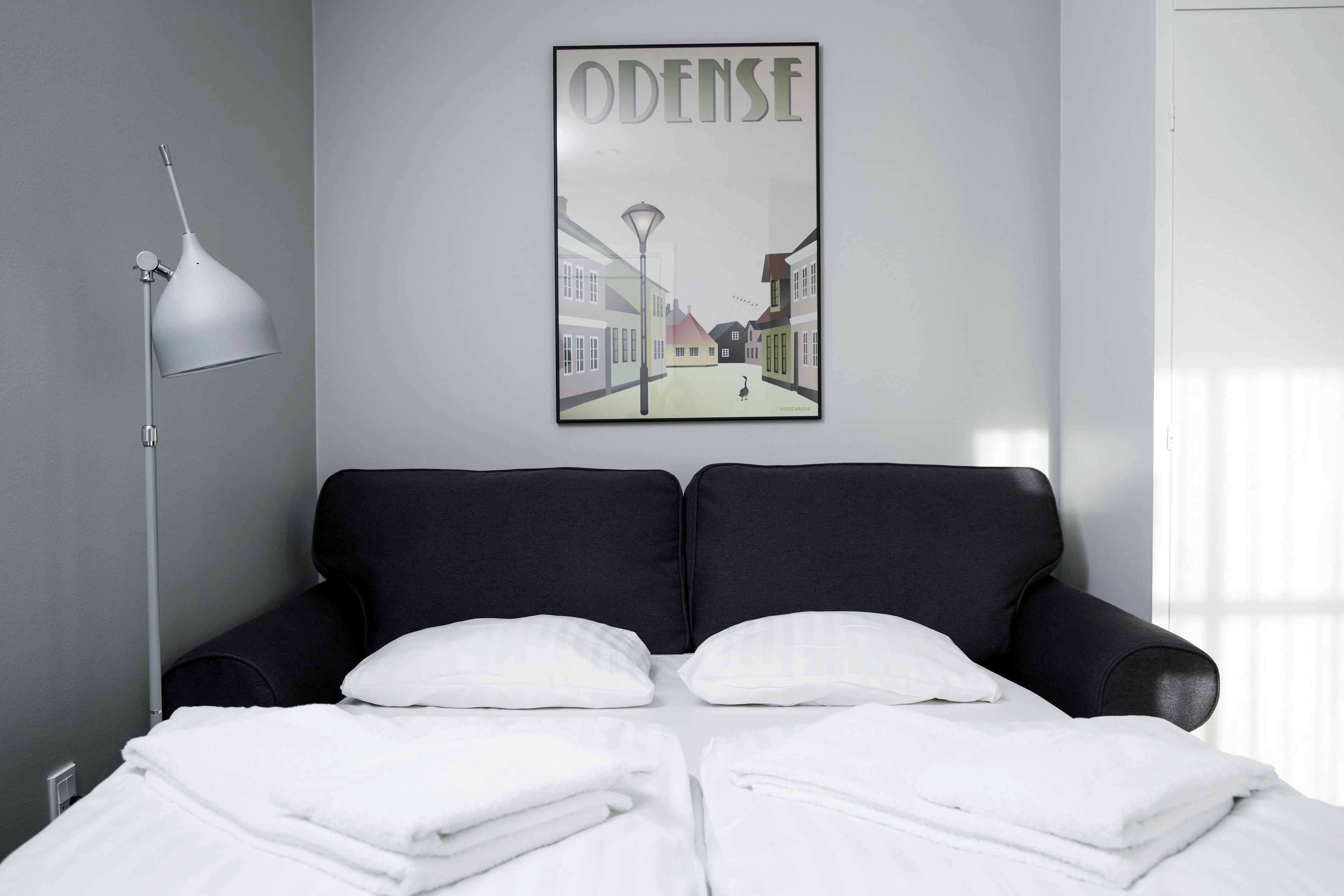 A modern bedroom with a black sofa bed, white bedding, a wall-mounted lamp, and a poster of a street scene.