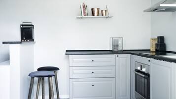 A modern kitchen with white cabinetry, a black countertop, and a built-in oven. There are two stools and a shelf with books and decorative items.