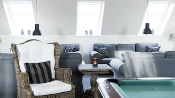 Een moderne woonkamer met een pooltafel, grijze banken, een rieten fauteuil en twee ramen met potplanten.