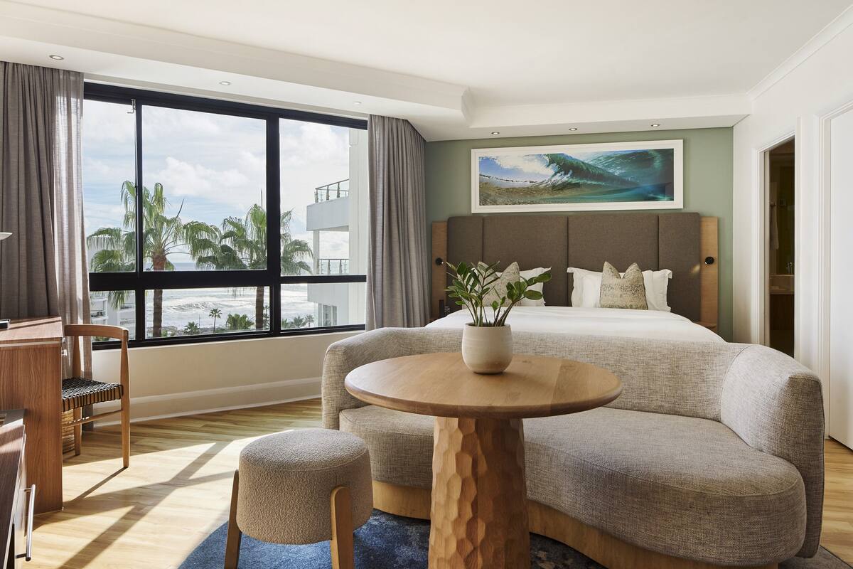 Ein Hotelzimmer mit einem großen Bett, einem runden Holztisch, einem Stuhl, einem Schreibtisch und einem großen Fenster mit Blick auf Palmen und Gebäude.