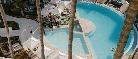 A rooftop pool area with lounge chairs and umbrellas, surrounded by palm trees and a multi-story building.