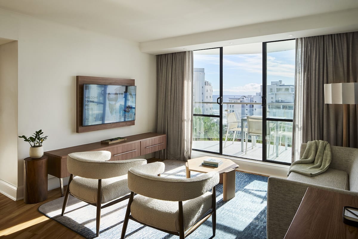 Ein modernes Hotelzimmer mit großem Fenster, Flachbildfernseher, einer Sitzecke, zwei Sesseln, einem kleinen Tisch und einem Schreibtisch mit Pflanze.