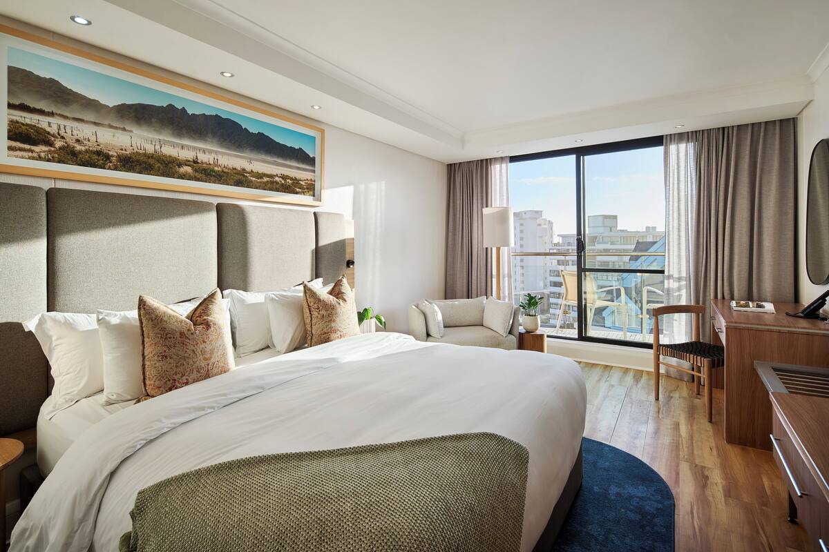 Ein Hotelzimmer mit einem großen Bett, einem Schreibtisch, einem Stuhl, einer Schreibtischlampe und Blick auf die Stadt durch große Fenster.