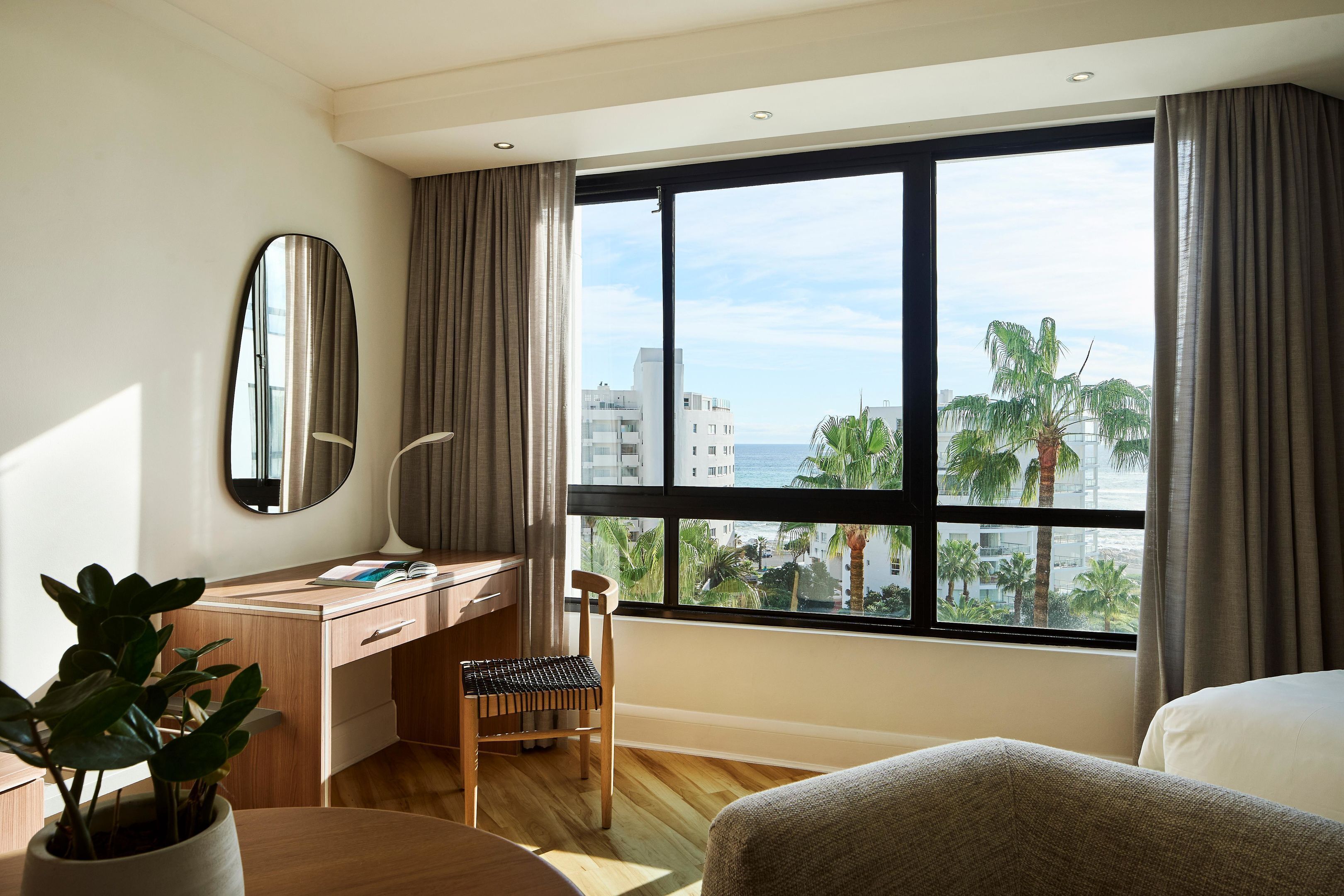 A hotel room with a large window, a desk, a chair, a bed, and a potted plant.