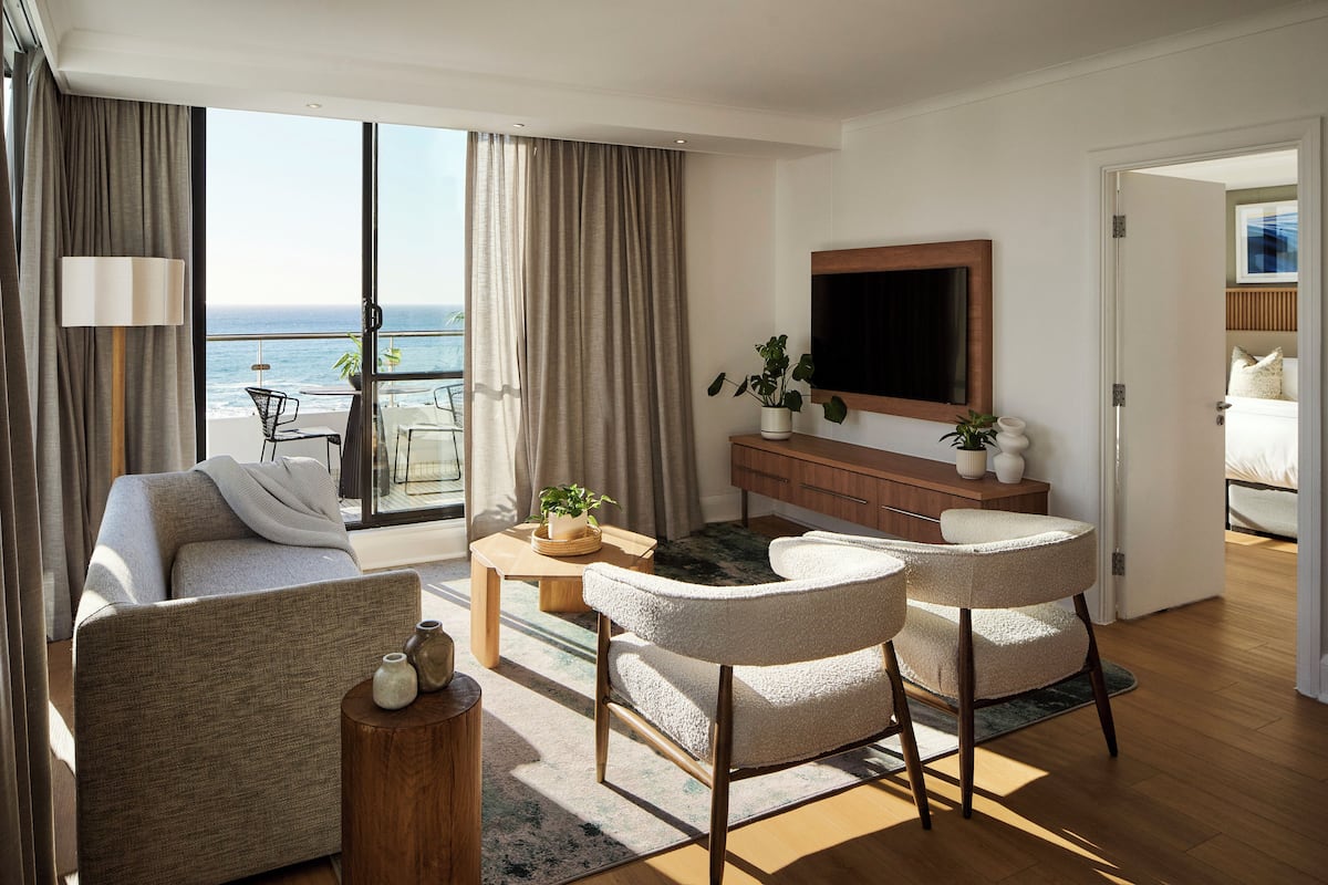 Ein modernes Wohnzimmer mit einer Couch, zwei Sesseln, einem Couchtisch und einem Fernseher. Es gibt einen Balkon mit Blick aufs Meer.