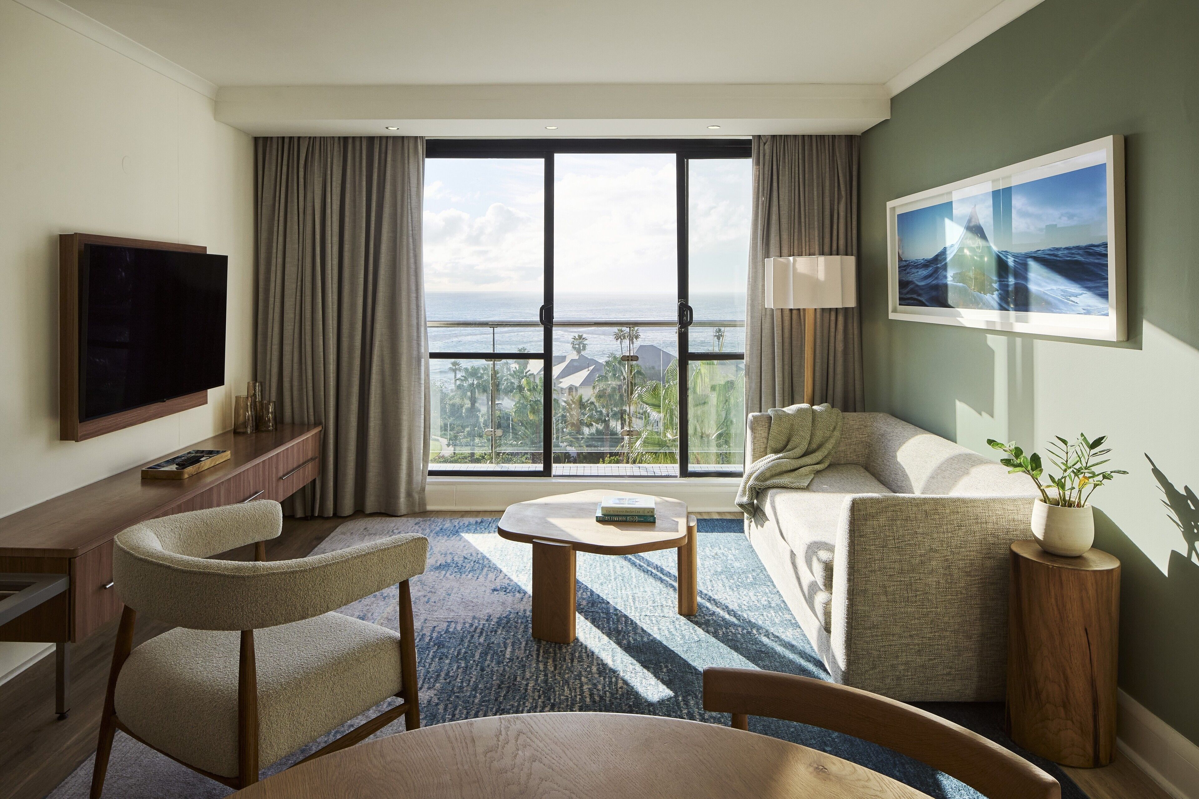 Een moderne hotelkamer met een groot raam, een flatscreen-tv, een bank, een eettafel en een klein bijzettafeltje met een plant.