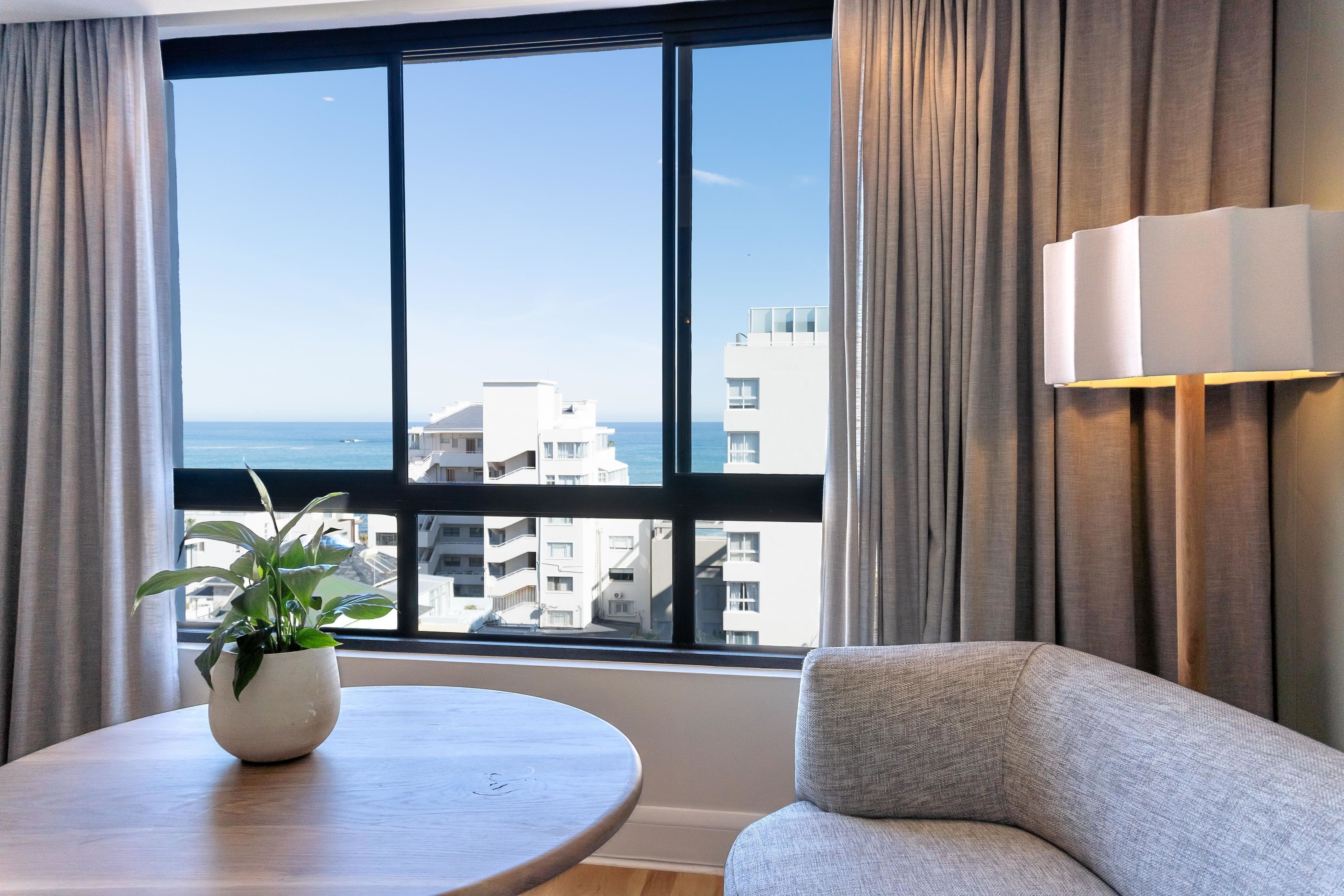 A room with a large window, a round table with a potted plant, a sofa, and a wall-mounted lamp.
