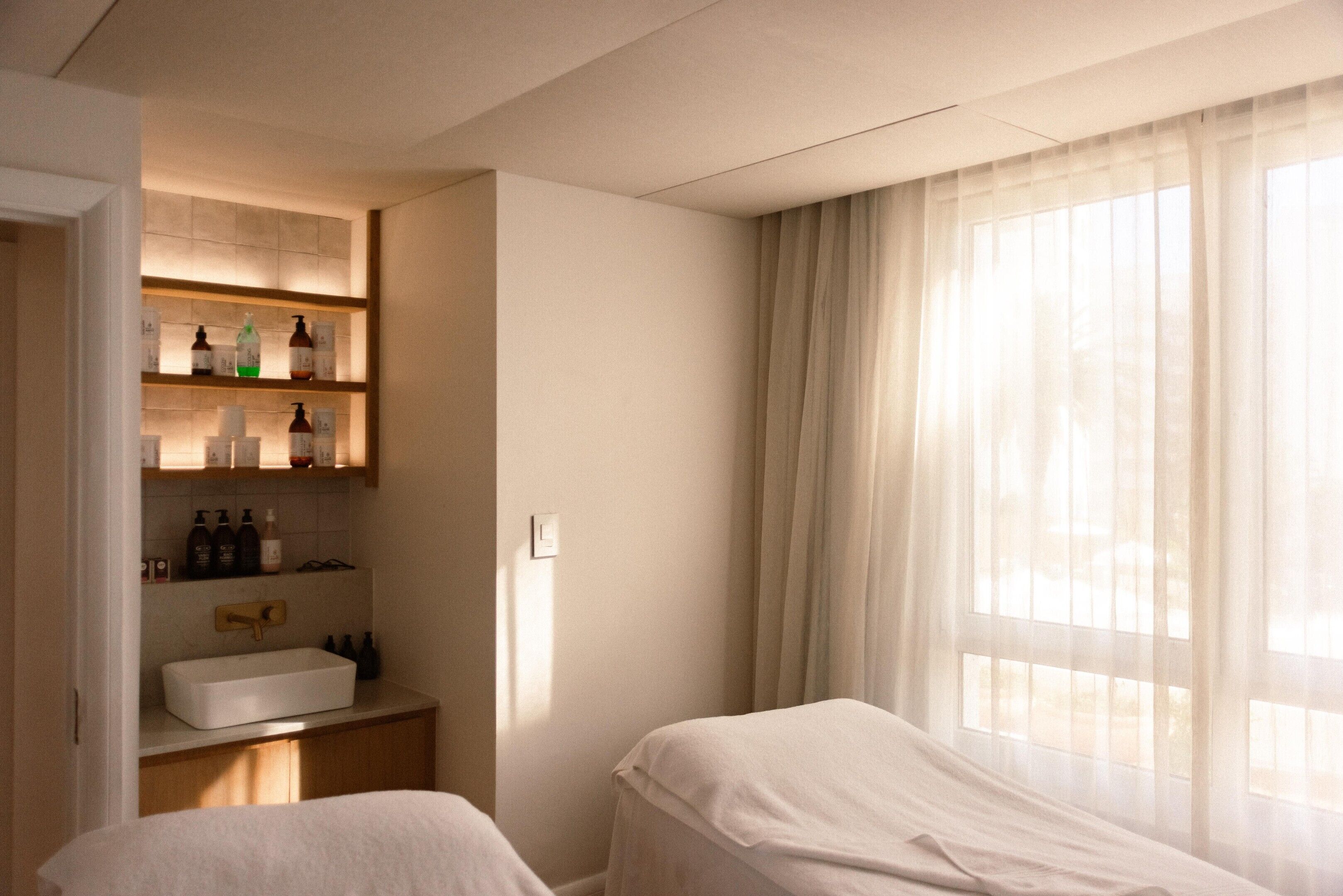 Et hotelværelse med indbygget hylde, toiletartikler, et natbord og et stort vindue med let gardin.
