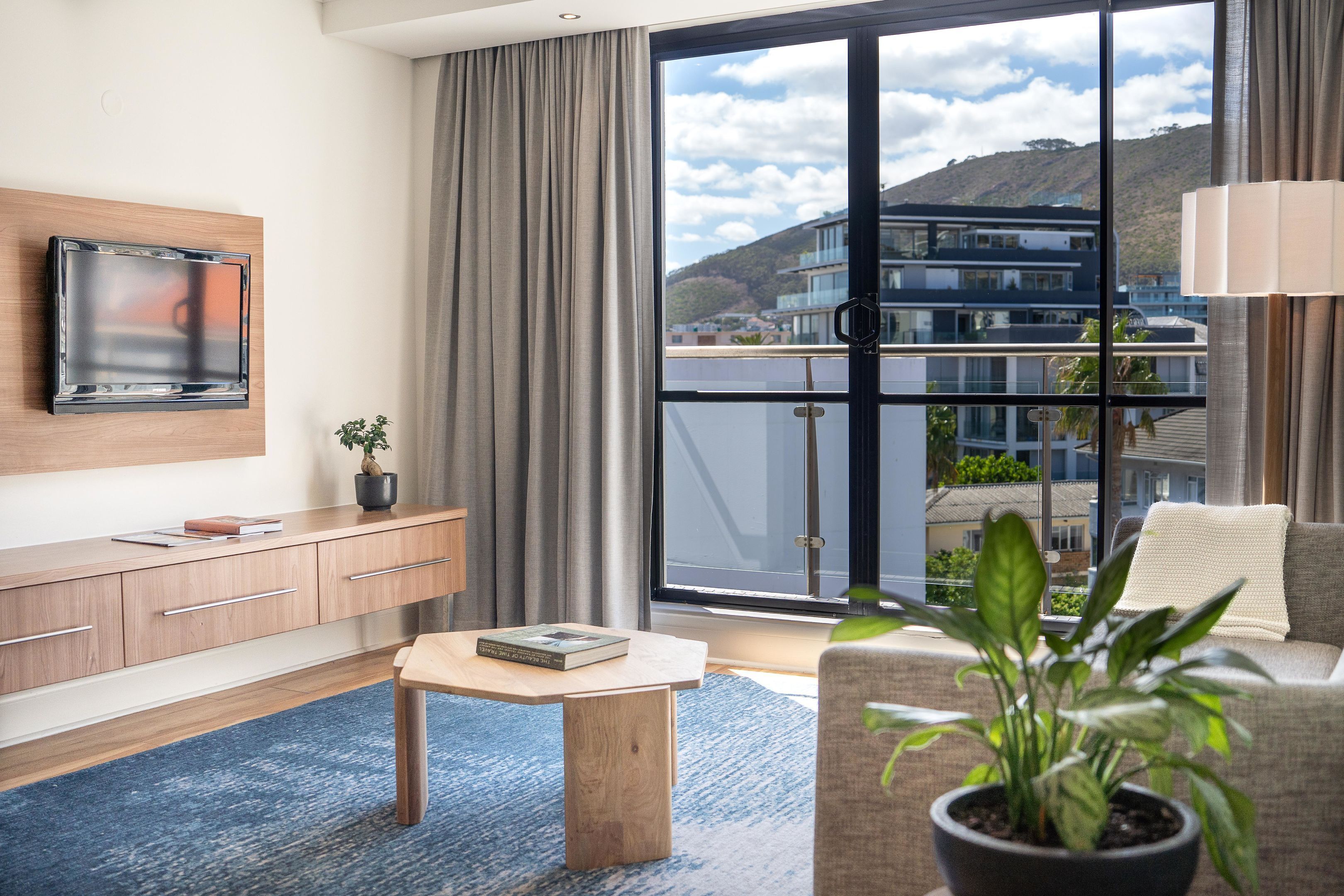 A modern hotel room with a large window, a flat-screen TV mounted on the wall, a wooden desk, and a potted plant.