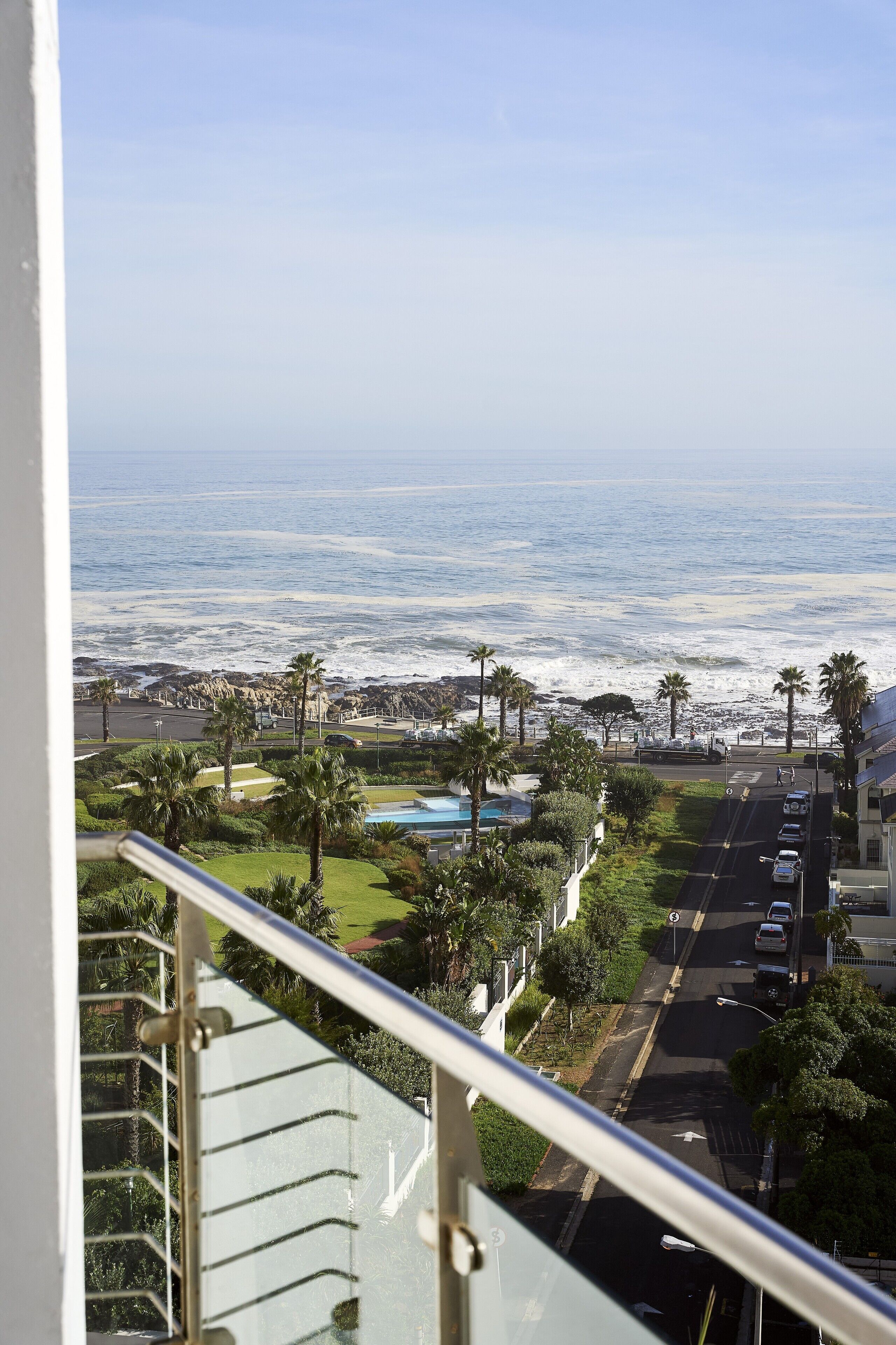 A coastal view from a balcony overlooking a street with palm trees, a swimming pool, and the ocean.