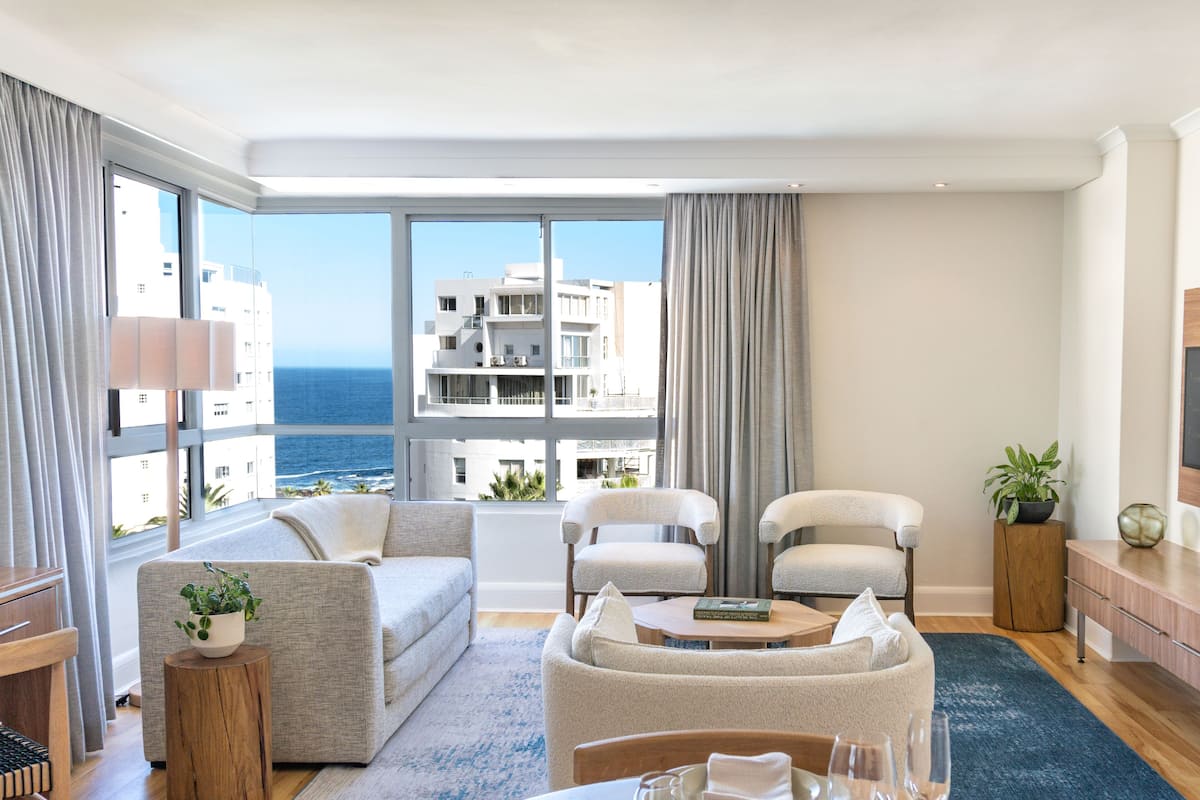 A modern living room with a sofa, two armchairs, a coffee table, and a dining table set for two. Large windows offer a view of the sea and buildings.