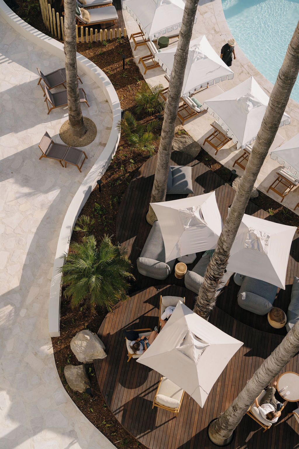 An outdoor lounge area with multiple seating options, including lounge chairs and umbrellas, surrounded by palm trees and a pool.