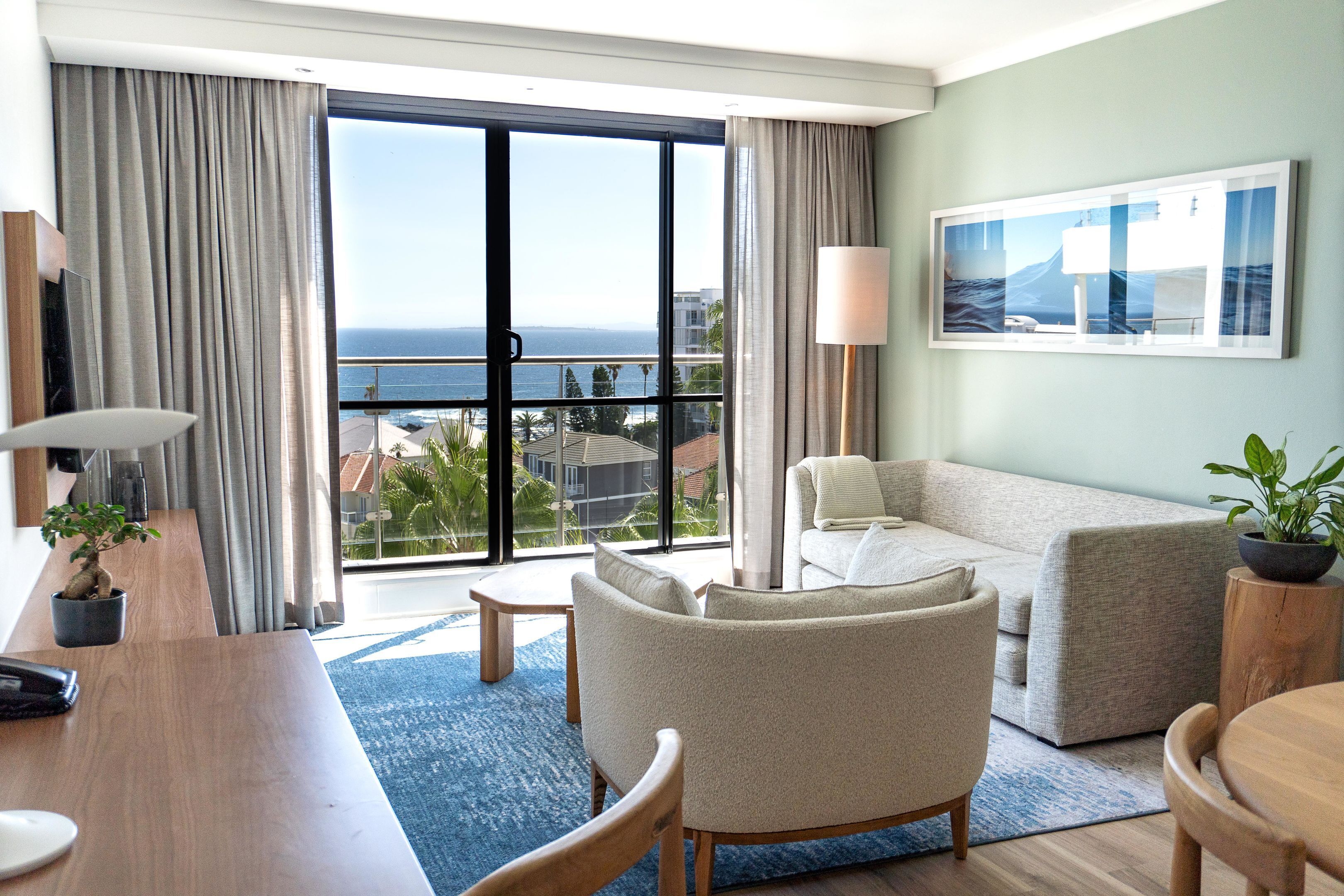 A hotel room with a large window, a sofa, a dining table, and a lamp.