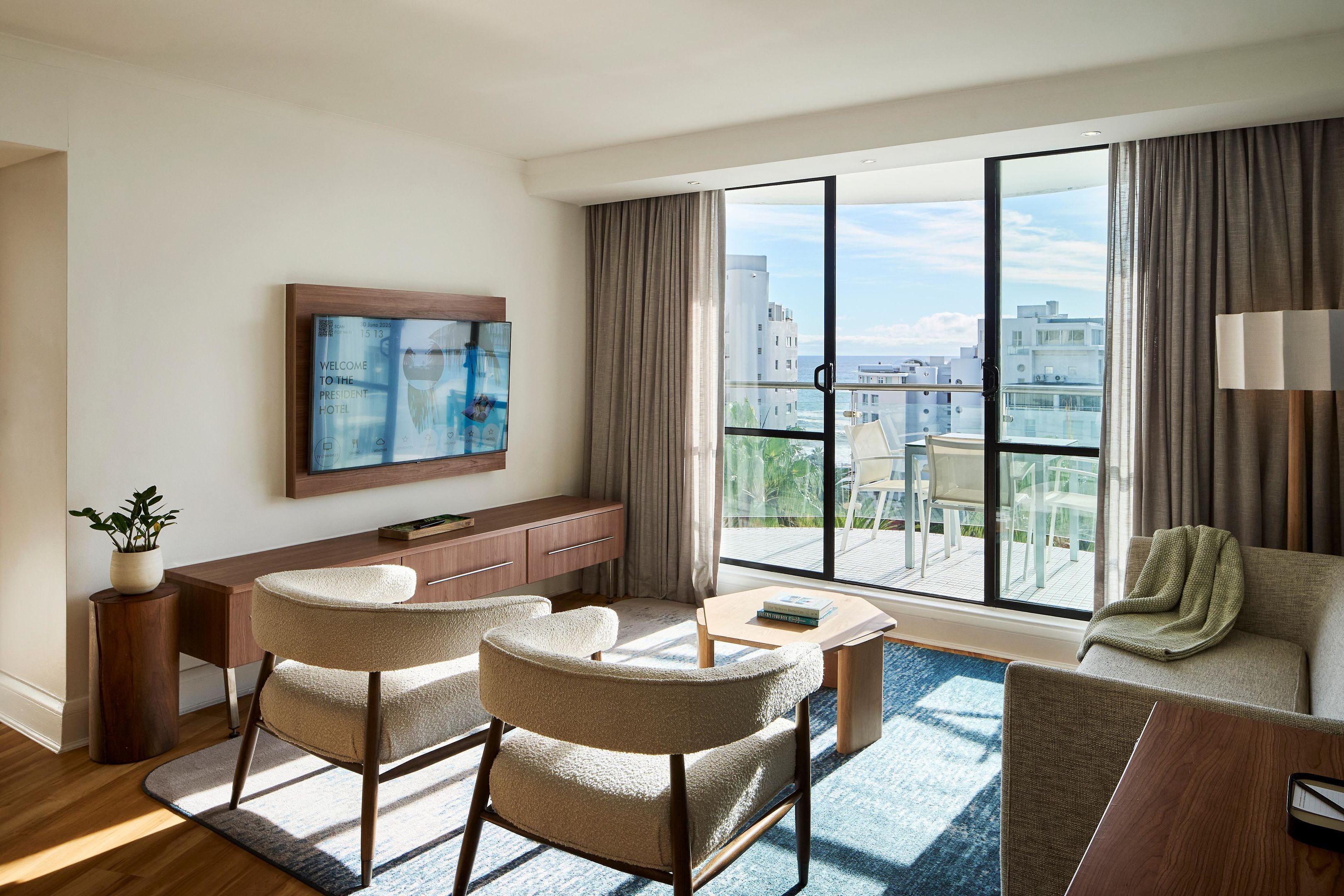 A modern hotel room with a large window, a flat-screen TV, a sofa, two armchairs, a wooden desk, and a small table.