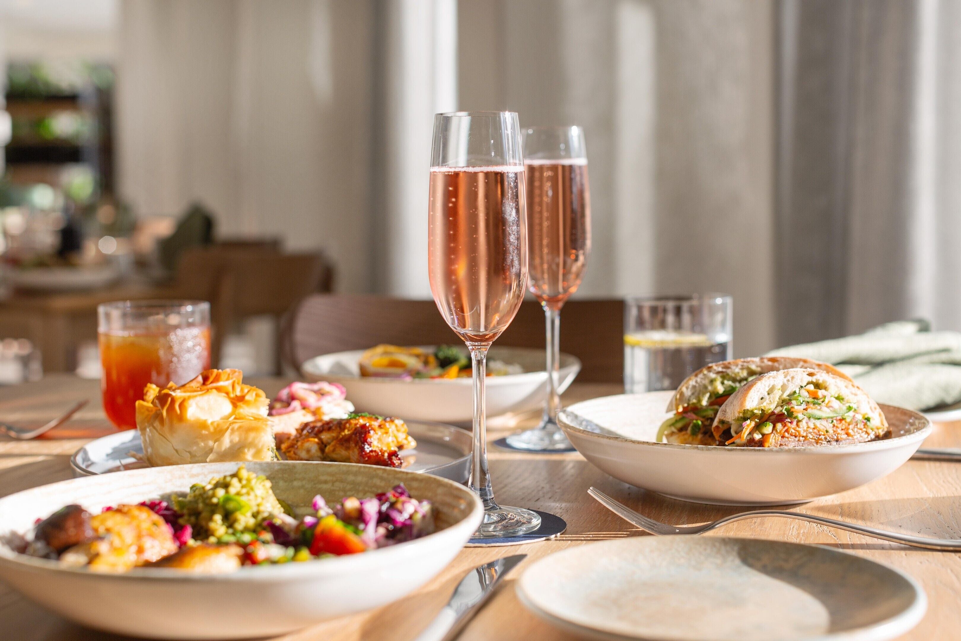 A dining table with various dishes, glasses of rosé wine, and a sandwich.