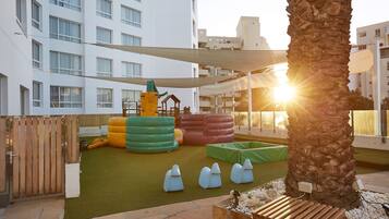 Children can play on the slide and climbing elements in this outdoor play area, surrounded by a palm tree and a wooden fence.