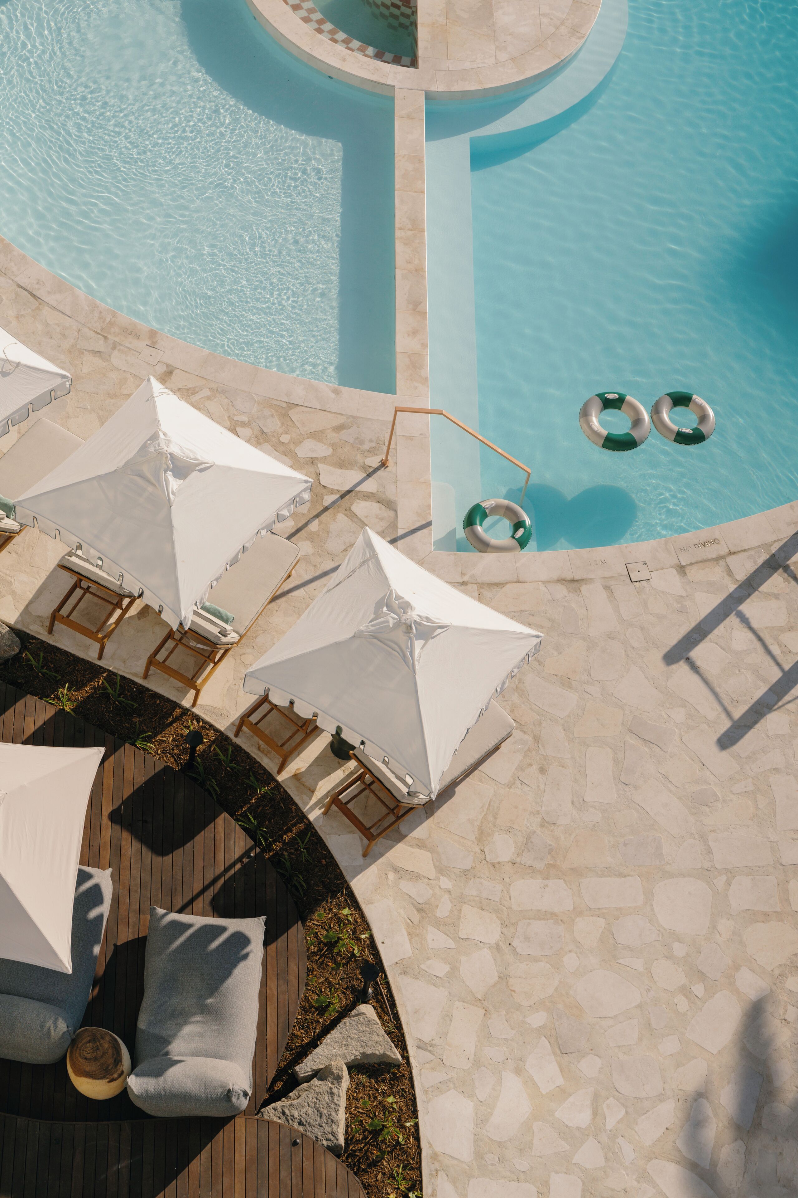 An aerial view of a pool area with lounge chairs and umbrellas, a stone deck, and a circular pool with a clear blue water.